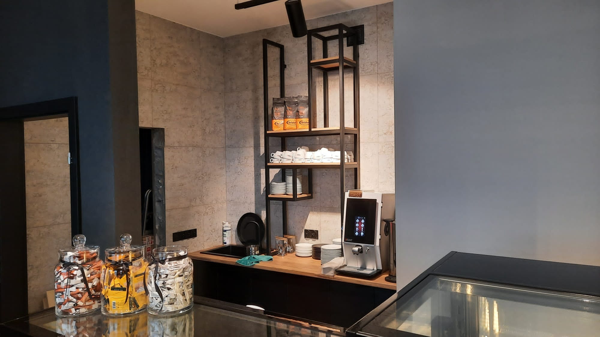 Nowoczesna strefa kawowa z ekspresami, czarną umywalką i półkami z kawą i filiżankami na tle szarej ściany z płytek.