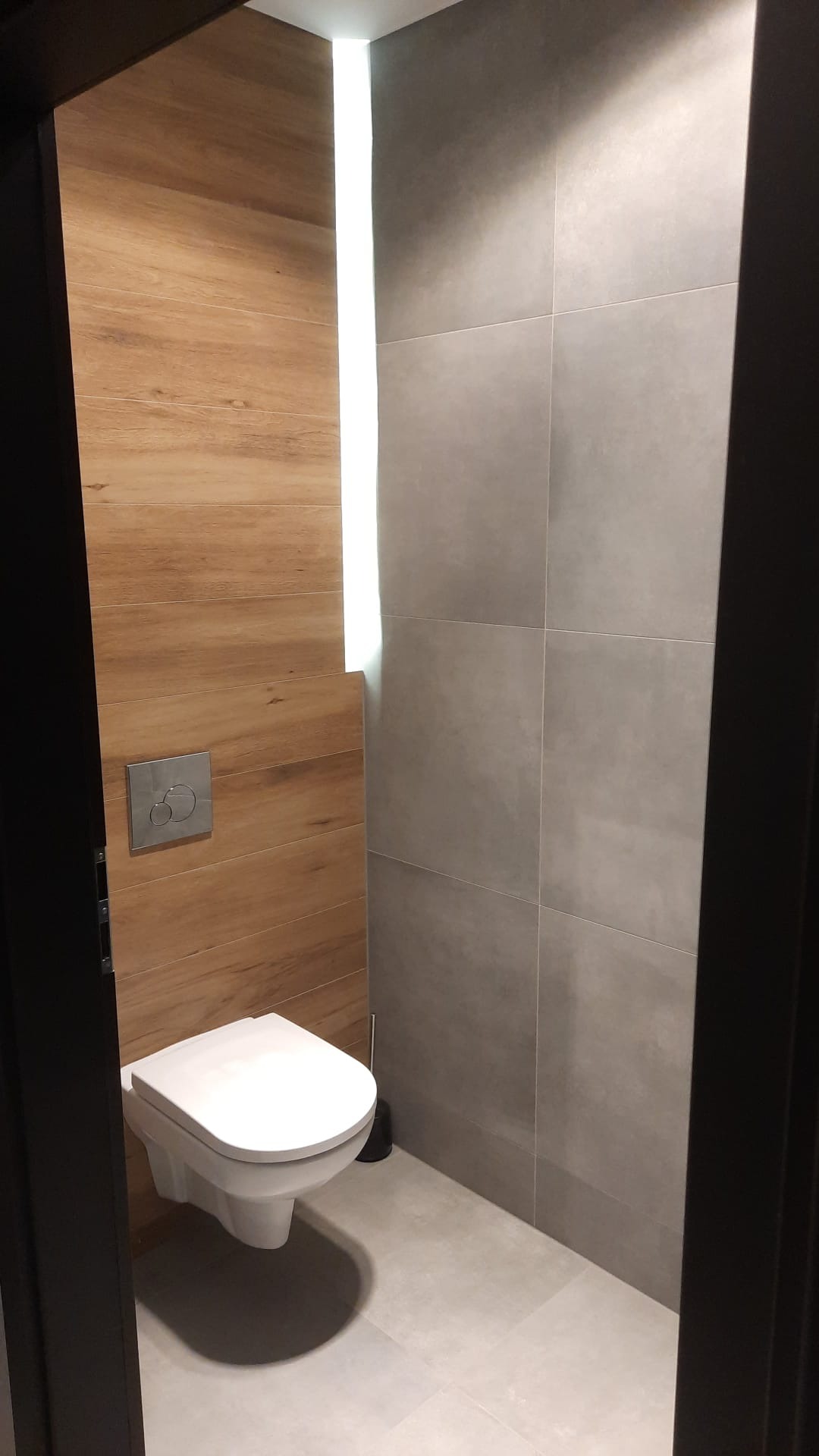Nowoczesna toaleta podwieszana w łazience wykończonej płytkami imitującymi drewno i szarym gresem, z podłużnym oświetleniem LED w rogu.