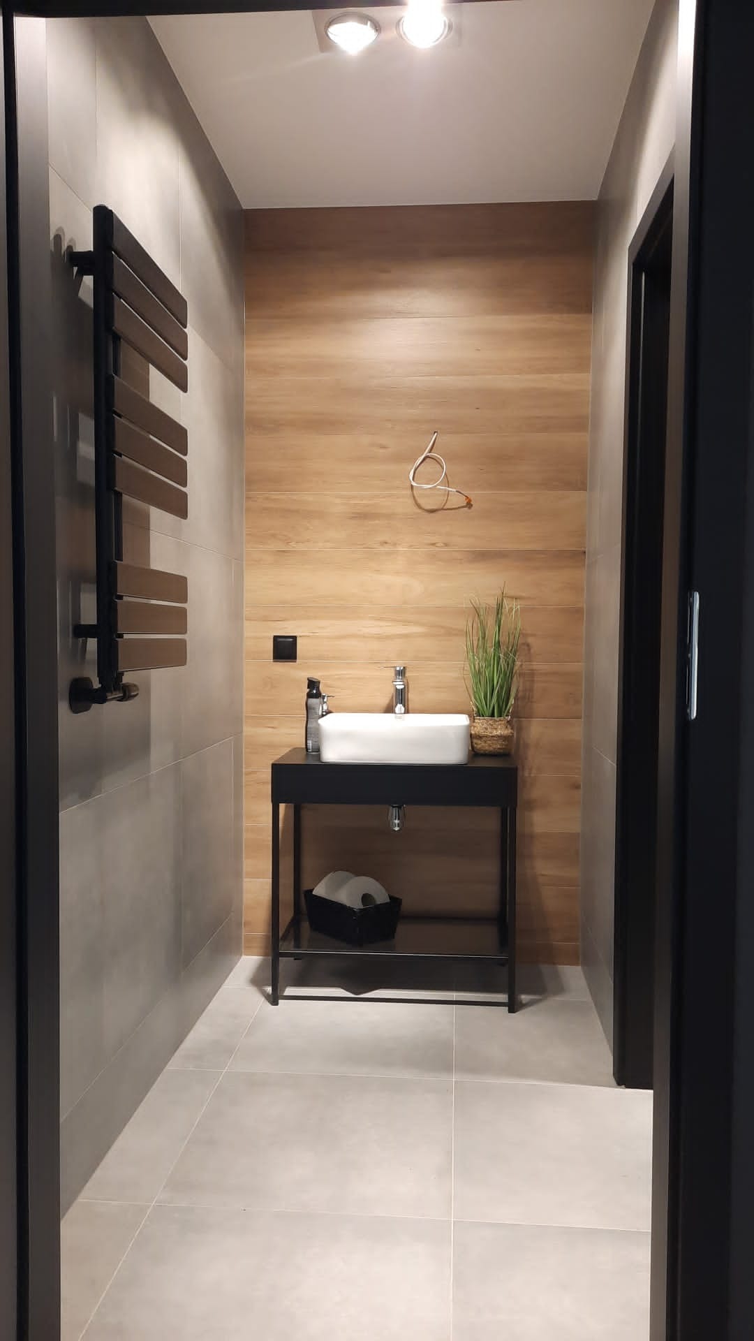 Nowoczesna łazienka z drewnianą ścianą, umywalką na czarnym stelażu, grzejnikiem drabinkowym i szarą glazurą.