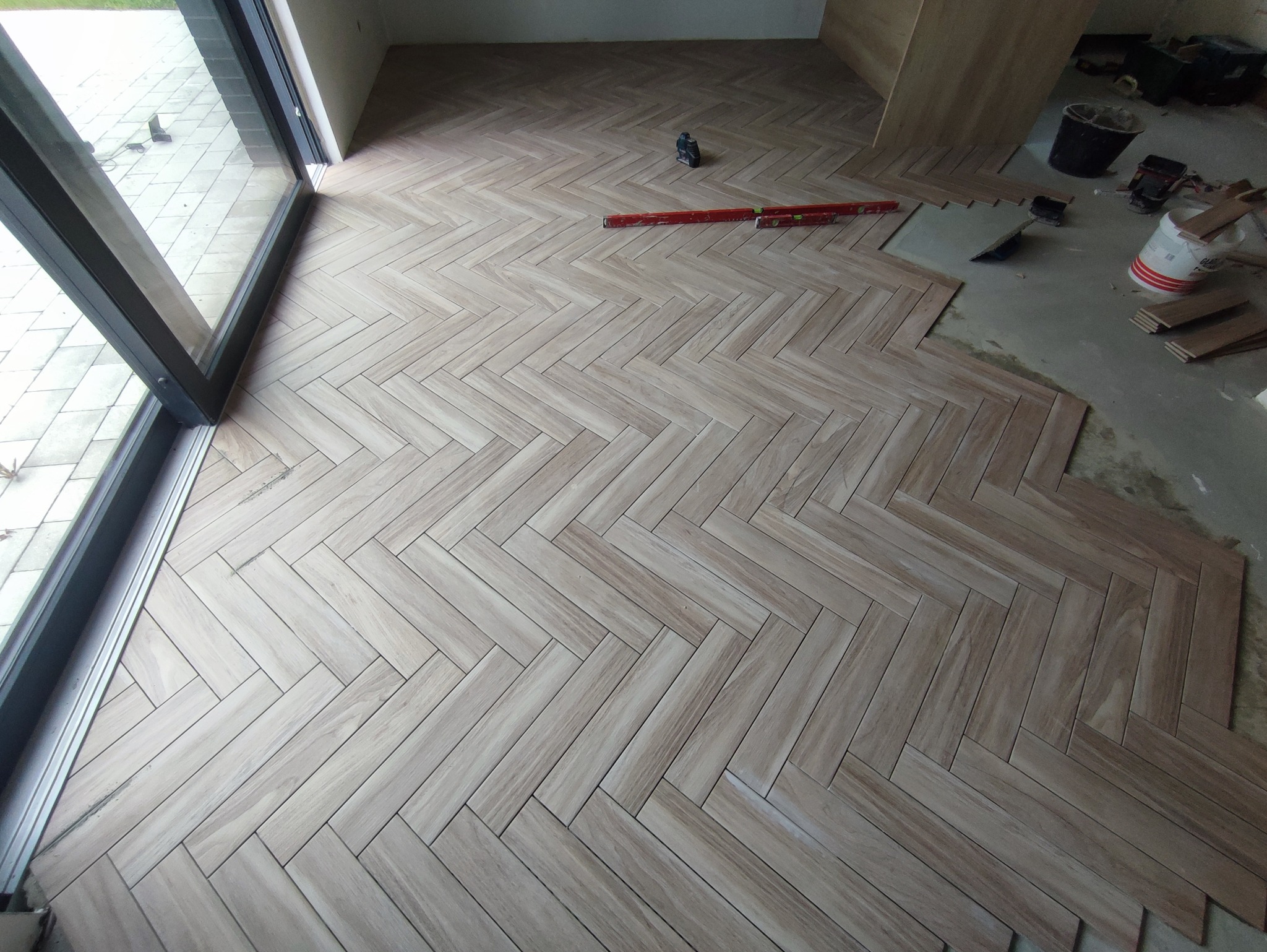 Układanie parkietu w jodełkę z płytek imitujących drewno, widoczne poziomice, wiadro z klejem i nieukończony fragment posadzki przy ścianie.