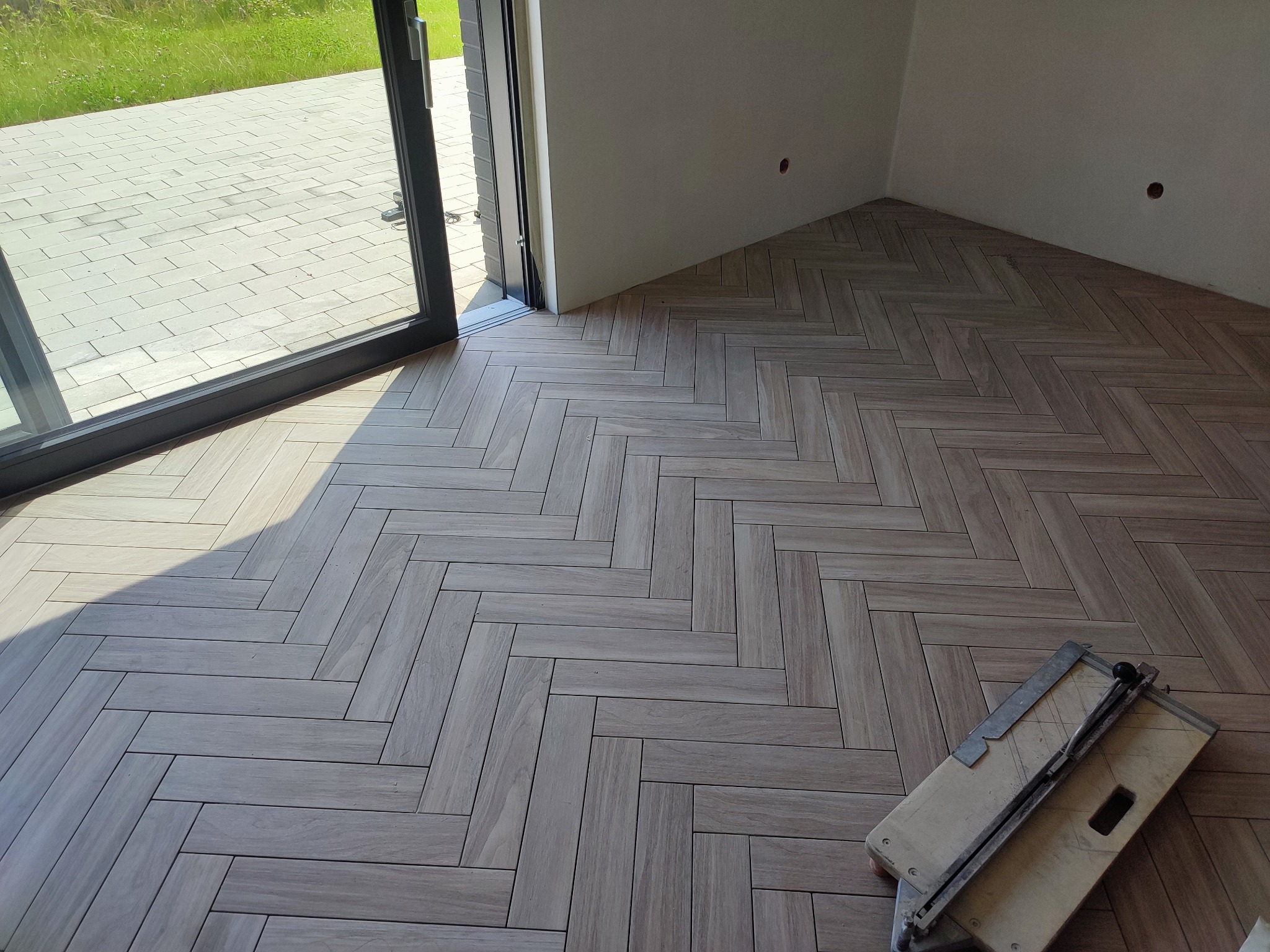 Ułożona podłoga z płytek imitujących drewno w jodełkę, widoczny fragment okna balkonowego i narzędzie do cięcia płytek.