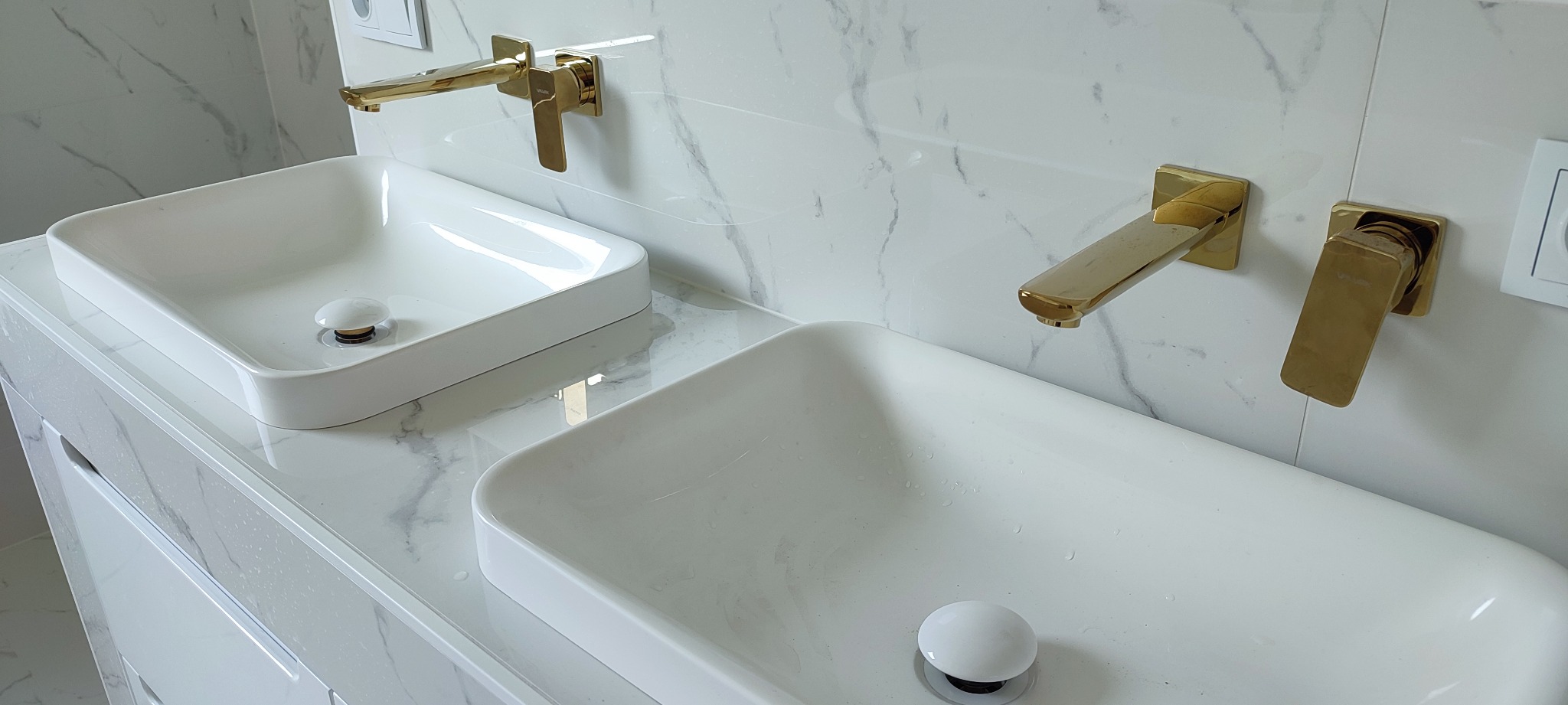 Dwie białe, prostokątne umywalki nablatowe z złotymi bateriami na tle ściany z marmurowym wzorem w nowoczesnej łazience.