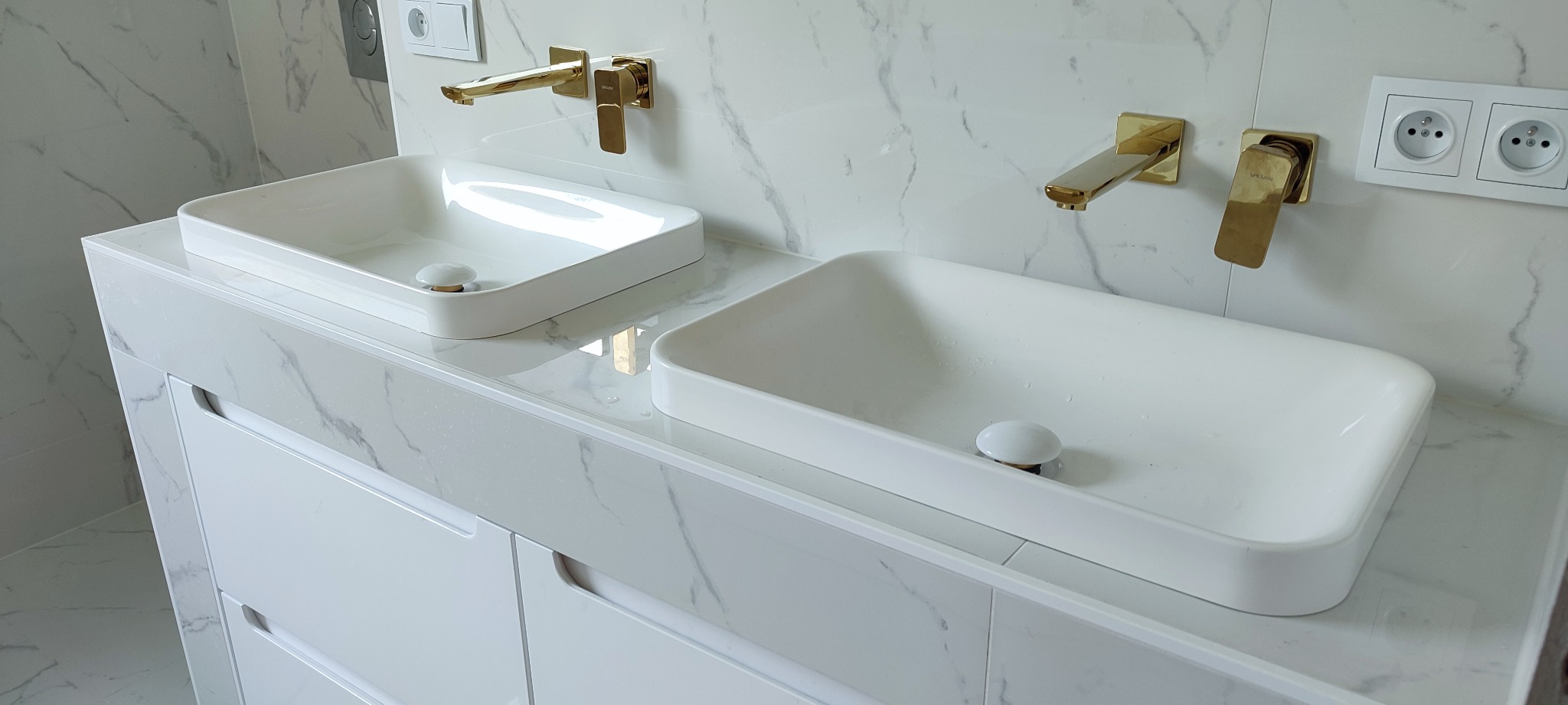 Biała łazienka z dwoma umywalkami nablatowymi, złotą armaturą i blatem imitującym marmur, widoczne gniazdka elektryczne.