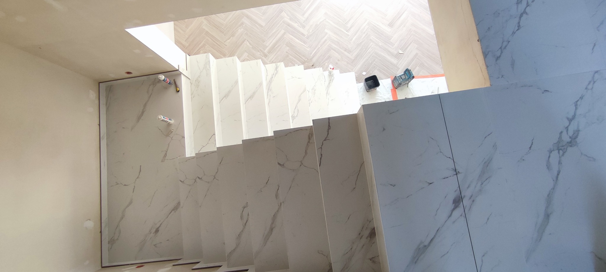 Widok z góry na schody wyłożone płytkami imitującymi marmur, z fragmentem parkietu u góry i narzędziami budowlanymi w tle, ukazujący etap prac wykończeniowych.