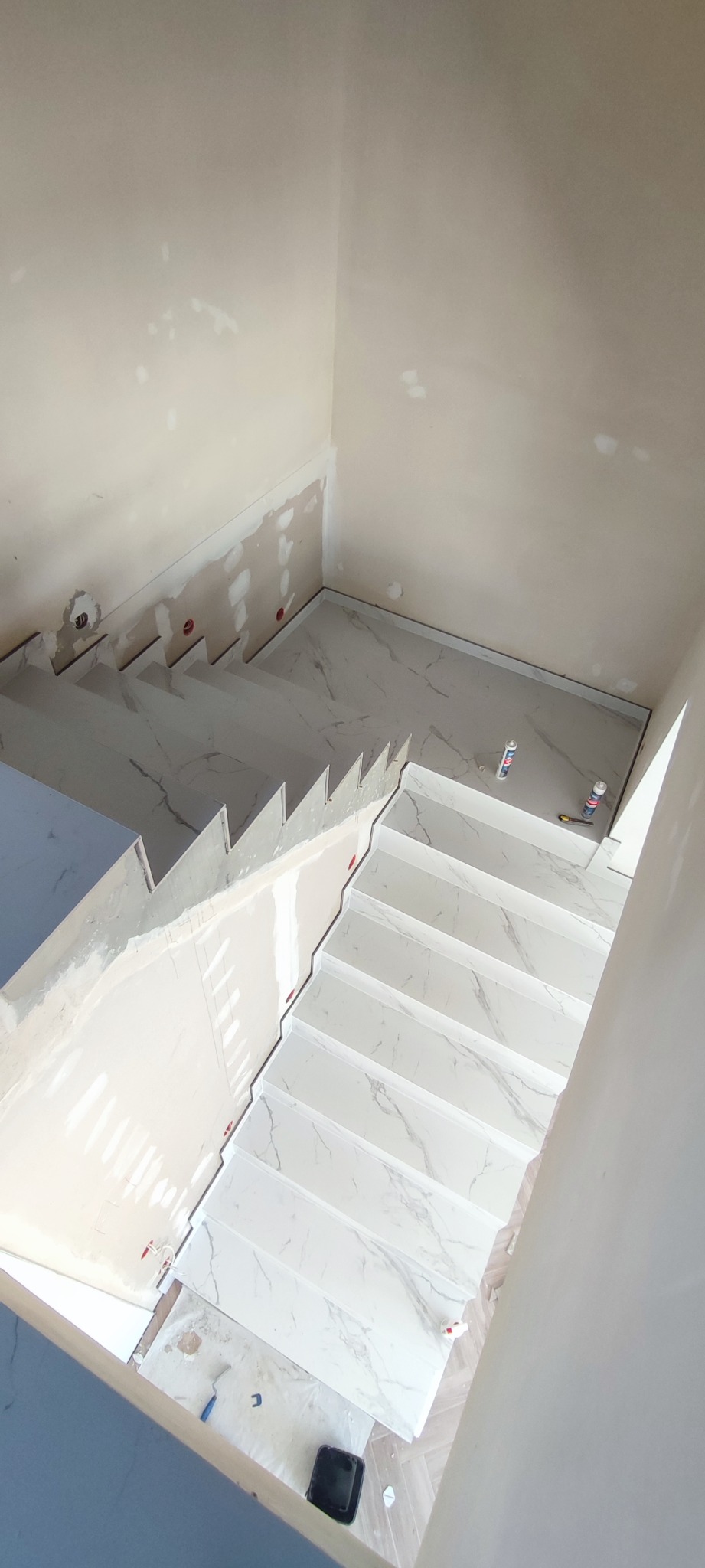 Wnętrze w trakcie wykańczania: schody wyłożone białymi płytkami imitującymi marmur, ściany w stanie surowym z widocznymi śladami po szpachlowaniu, tuby z klejem i narzędzia na podłodze.