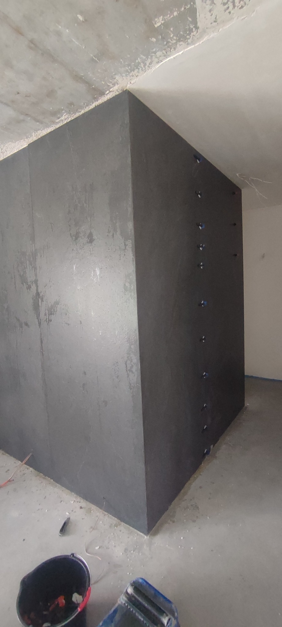 Narożnik łazienki z ciemnymi płytami, zamocowanymi na kołki rozporowe, widoczne niedokończone wykończenie sufitu i podłogi, wiadro i narzędzia na podłodze.