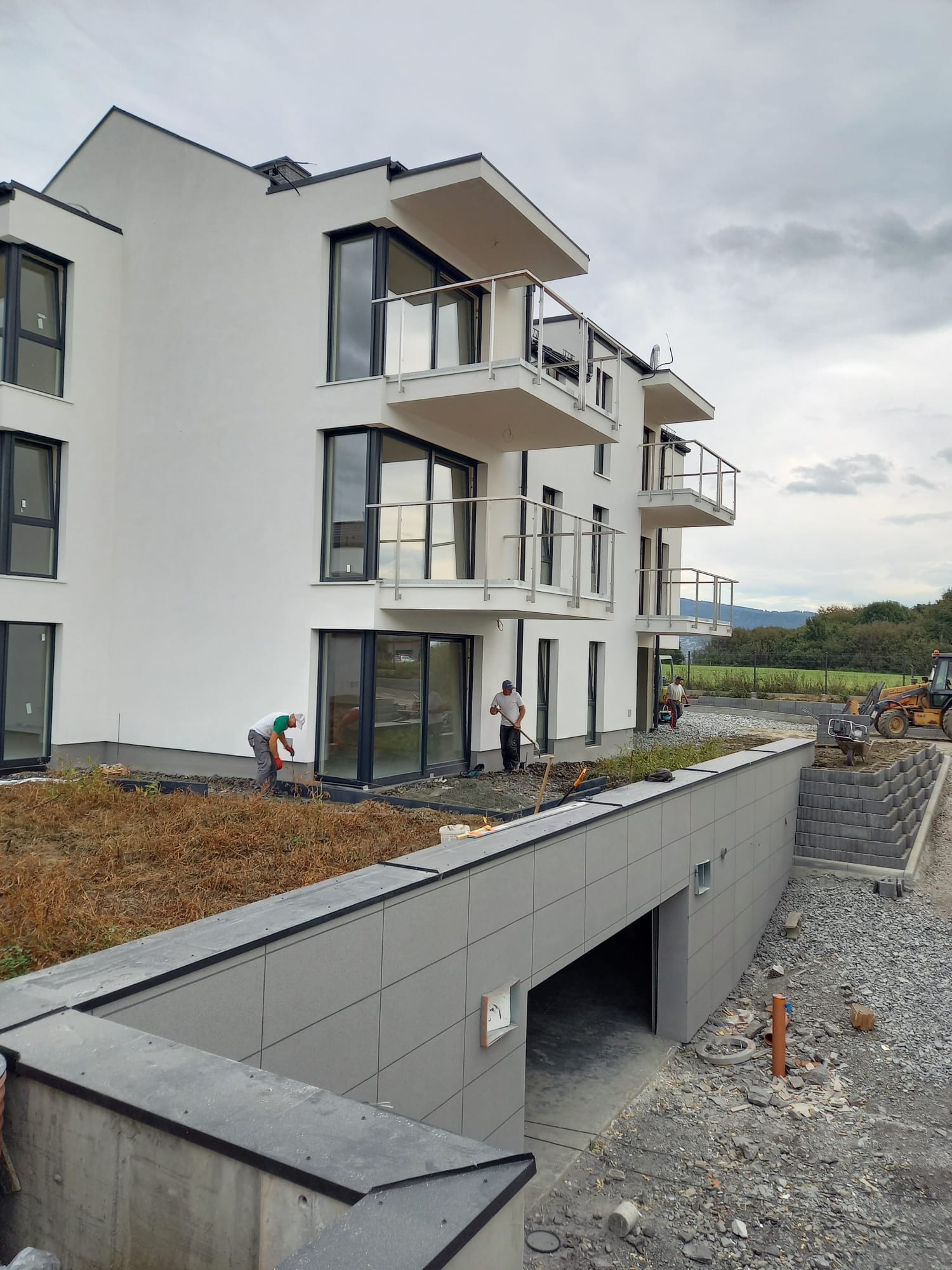 Nowoczesny budynek wielorodzinny w trakcie wykończenia, z pracownikami porządkującymi teren wokół, widoczny wjazd do garażu podziemnego i traktor w tle.