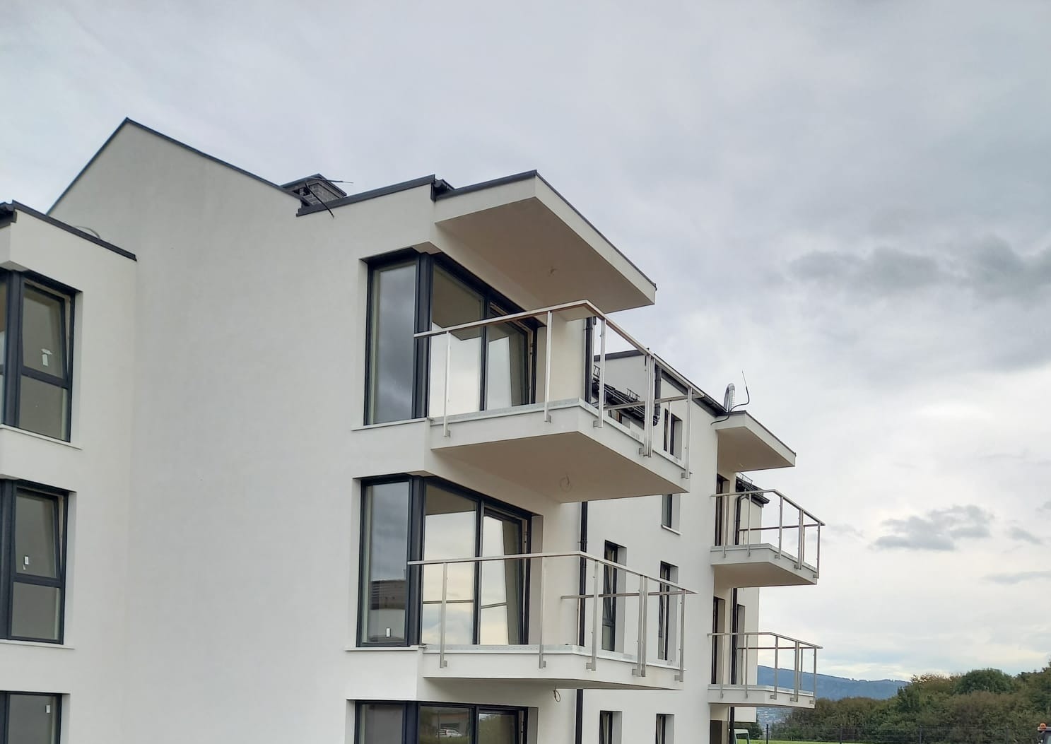 Nowoczesny budynek wielorodzinny z białymi elewacjami, balkonami z szklanymi balustradami i czarnymi ramami okiennymi, widziany pod pochmurnym niebem.