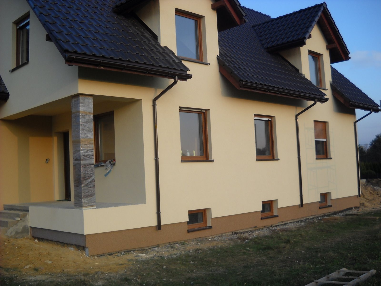 Jasna elewacja domu jednorodzinnego z ciemną dachówką, widoczne okna z brązowymi ramami, rynny, kolumna wejściowa z okładziną imitującą kamień oraz fragment terenu przed budynkiem.