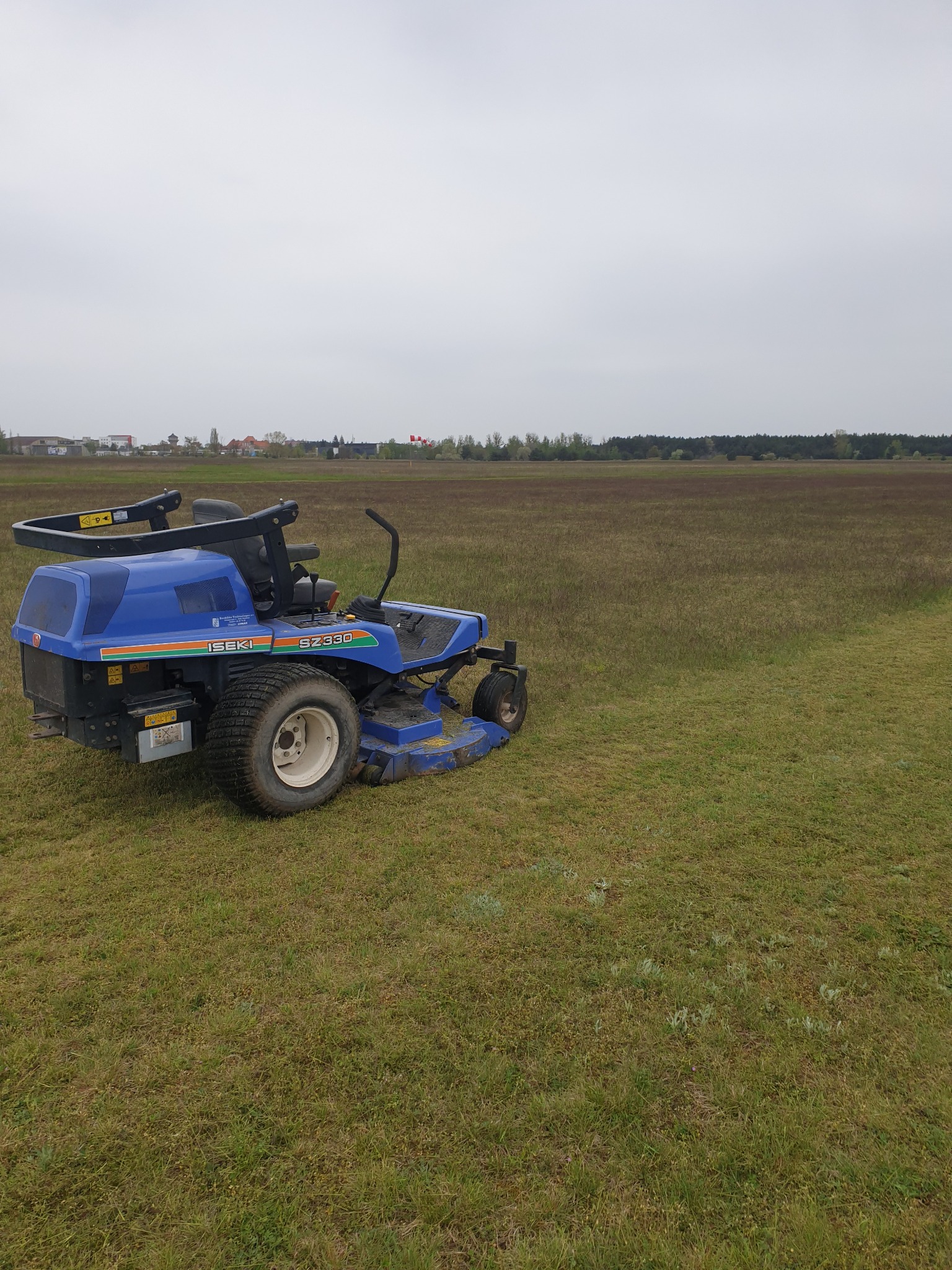 Niebieska kosiarka samojezdna Iseki SZ330 na nierównym, częściowo skoszonym trawniku, widoczne ślady cięcia, w tle zabudowania i las pod pochmurnym niebem.