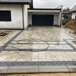 Nowa, częściowo ułożona nawierzchnia z kostki brukowej w odcieniach szarości i czerni przed domem z garażem, widoczny wąż ogrodowy i sterta ziemi.