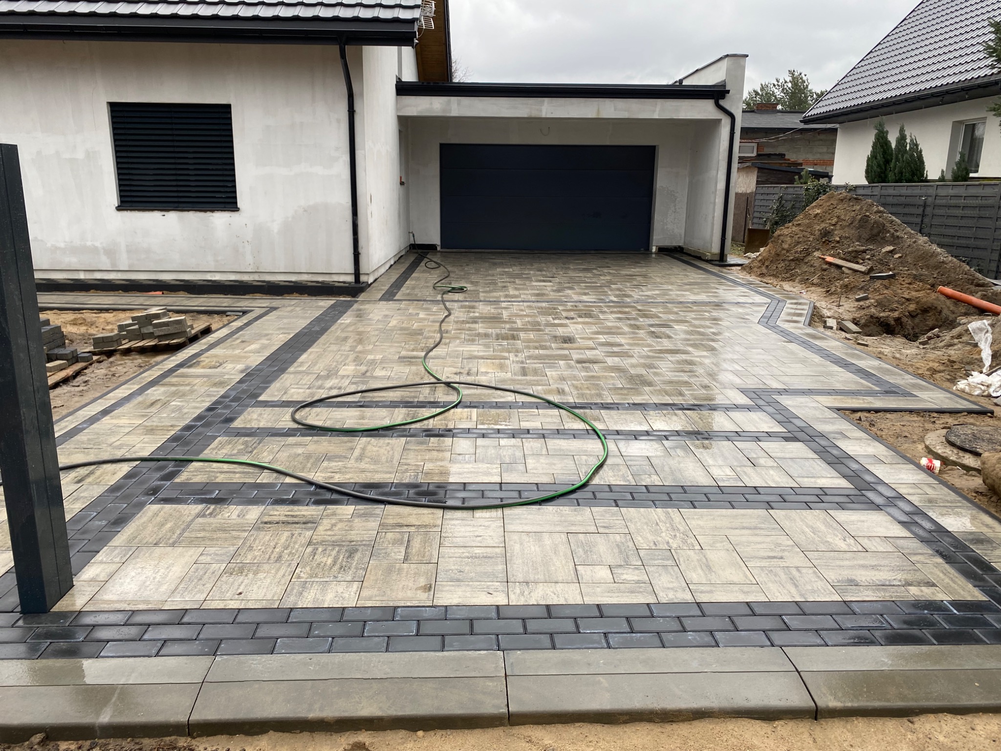 Nowa, częściowo ułożona nawierzchnia z kostki brukowej w odcieniach szarości i czerni przed domem z garażem, widoczny wąż ogrodowy i sterta ziemi.