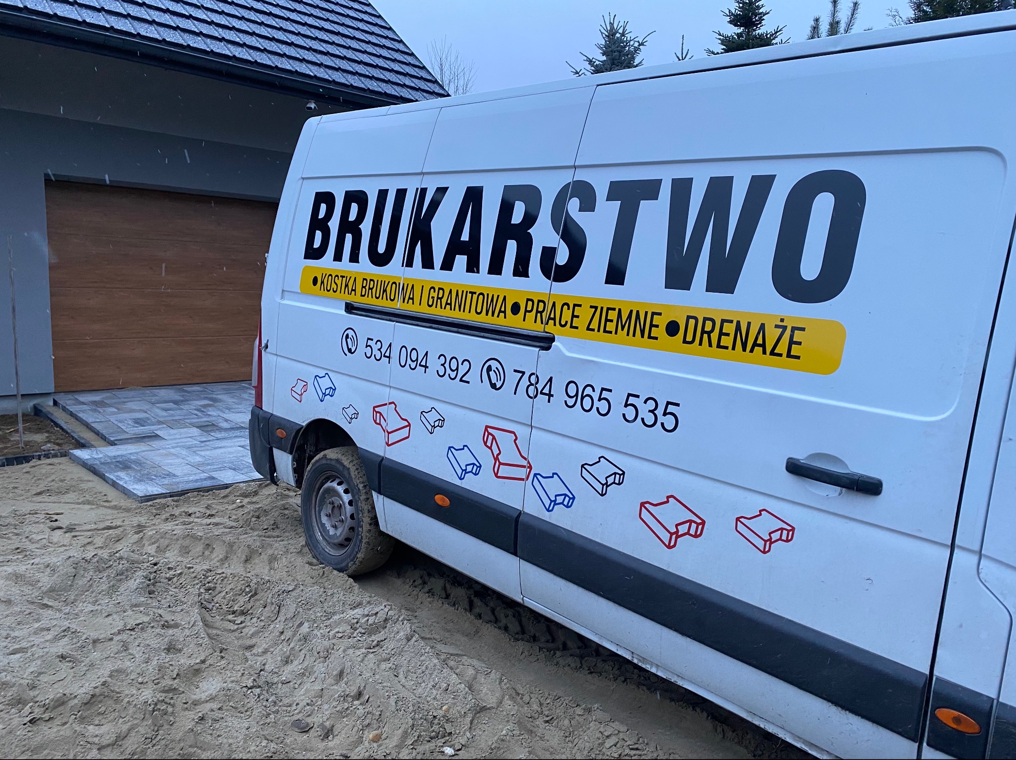 Biały samochód dostawczy z napisem 'BRUKARSTWO' i numerami telefonów, zaparkowany na piasku obok ułożonej częściowo kostki brukowej przed garażem.