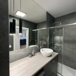 Nowoczesna łazienka w odcieniach szarości z umywalką nablatową, kabiną prysznicową ze szklanymi drzwiami przesuwnymi, podwieszanymi lampami cylindrycznymi i pralką ukrytą w szafce pod umywalką.