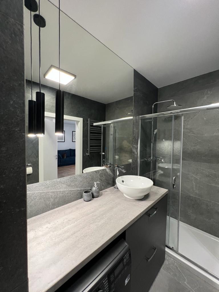 Nowoczesna łazienka w odcieniach szarości z umywalką nablatową, kabiną prysznicową ze szklanymi drzwiami przesuwnymi, podwieszanymi lampami cylindrycznymi i pralką ukrytą w szafce pod umywalką.