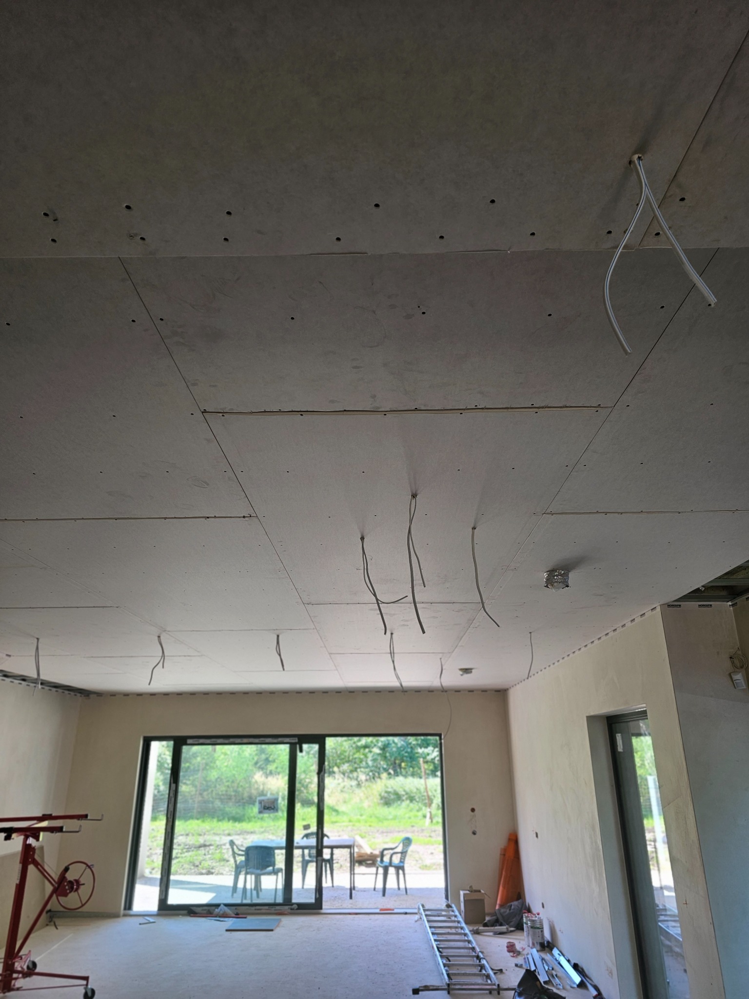 Montaż sufitu podwieszanego z płyt kartonowo-gipsowych, widoczne kable wystające z otworów, drabina i podnośnik do płyt w pomieszczeniu z dużym oknem tarasowym.