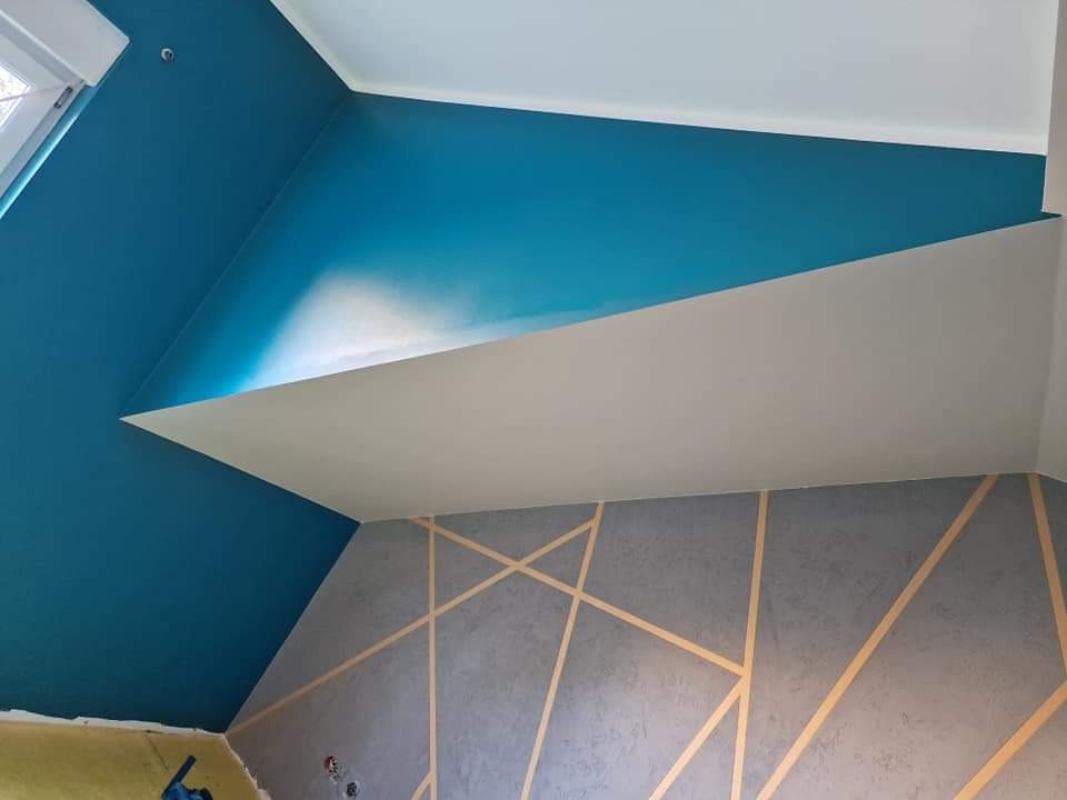 Ściana w pokoju na poddaszu pomalowana w kolorze turkusowym i szarym z geometrycznym wzorem z taśmy malarskiej. Widoczny fragment okna dachowego i niebieskie narzędzie malarskie na podłodze.