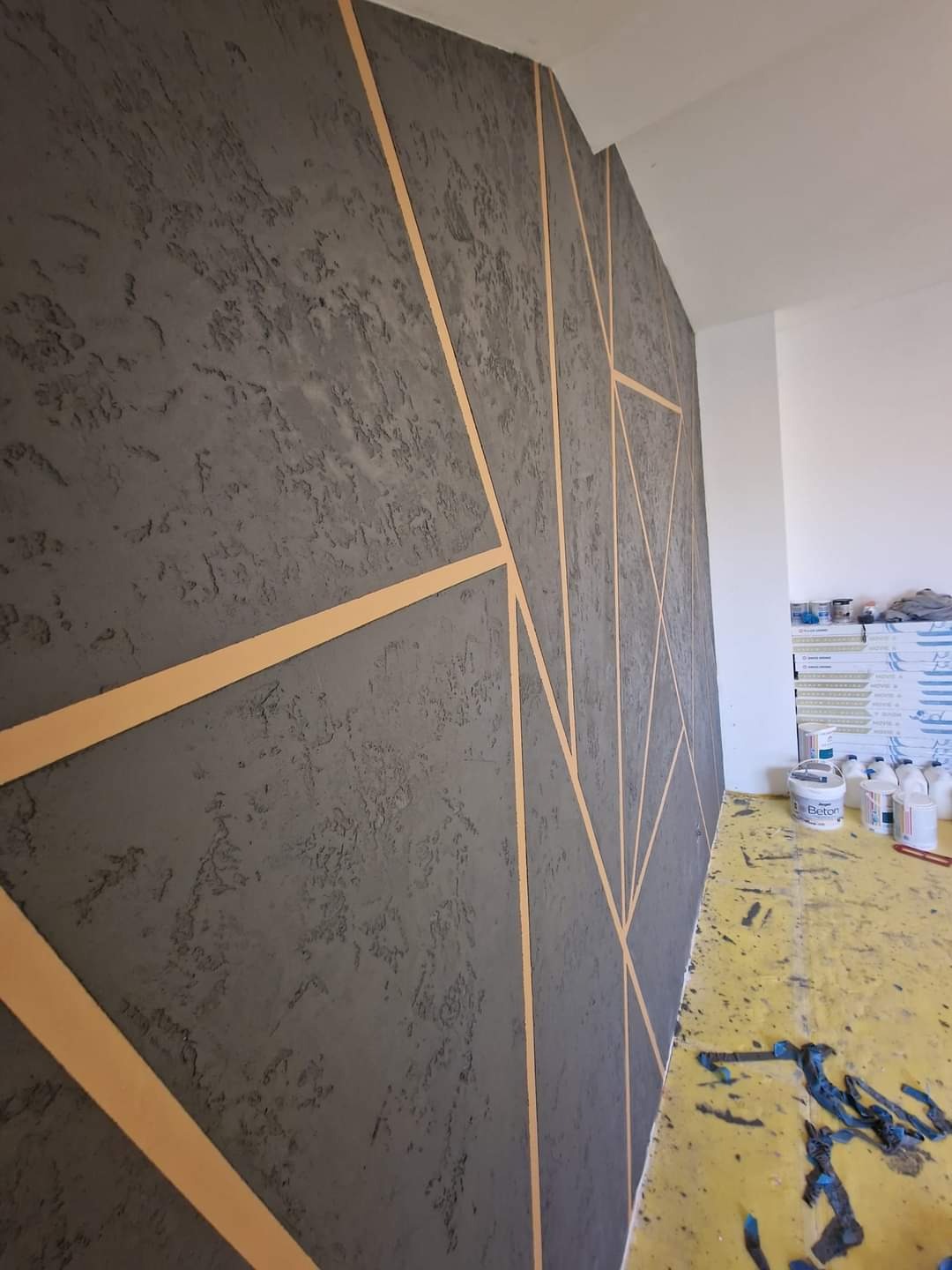 Dekoracyjna ściana z szarego betonu architektonicznego z geometrycznym wzorem z cienkich, złotych pasków, widok z boku, na podłodze ślady farby i pojemniki z materiałami.