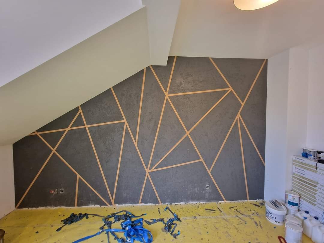 Ściana w pokoju na poddaszu pomalowana na szaro z geometrycznym wzorem z jasnych pasków, zabezpieczona folią podłoga, widoczne puszki z farbą i narzędzia.