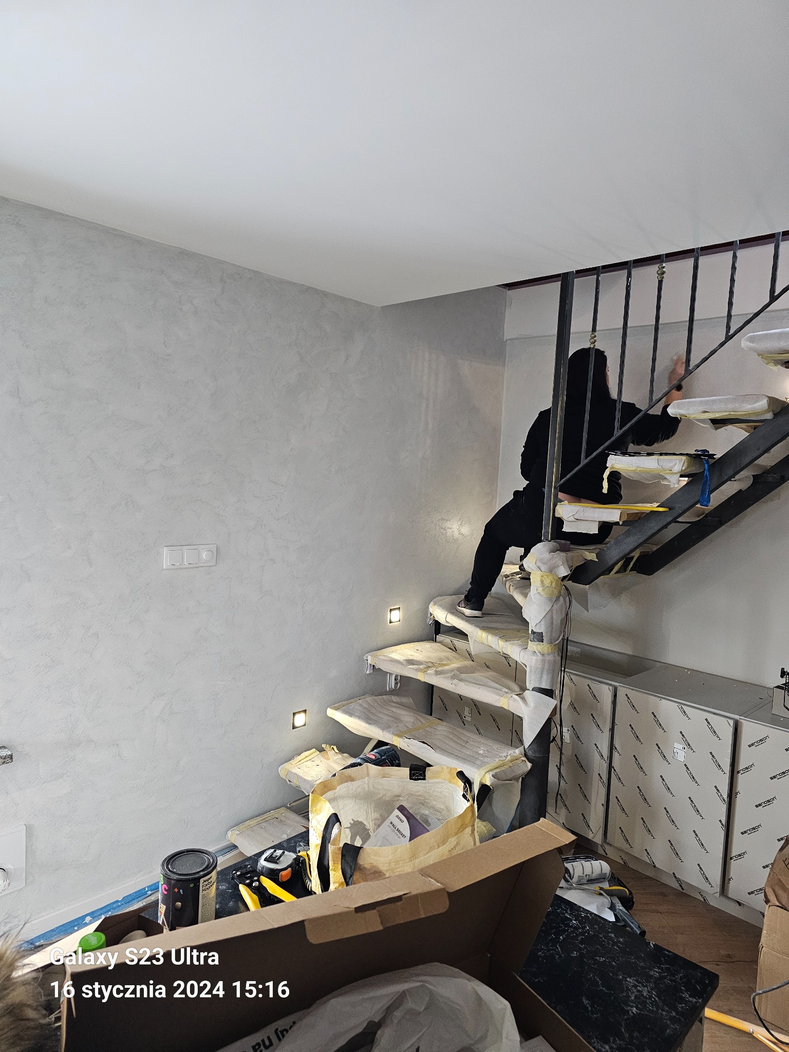 Pracownik w czarnym stroju wygładza ścianę o szarej, nieregularnej fakturze obok schodów zabezpieczonych folią malarską, widoczne narzędzia i puszka z farbą na podłodze.