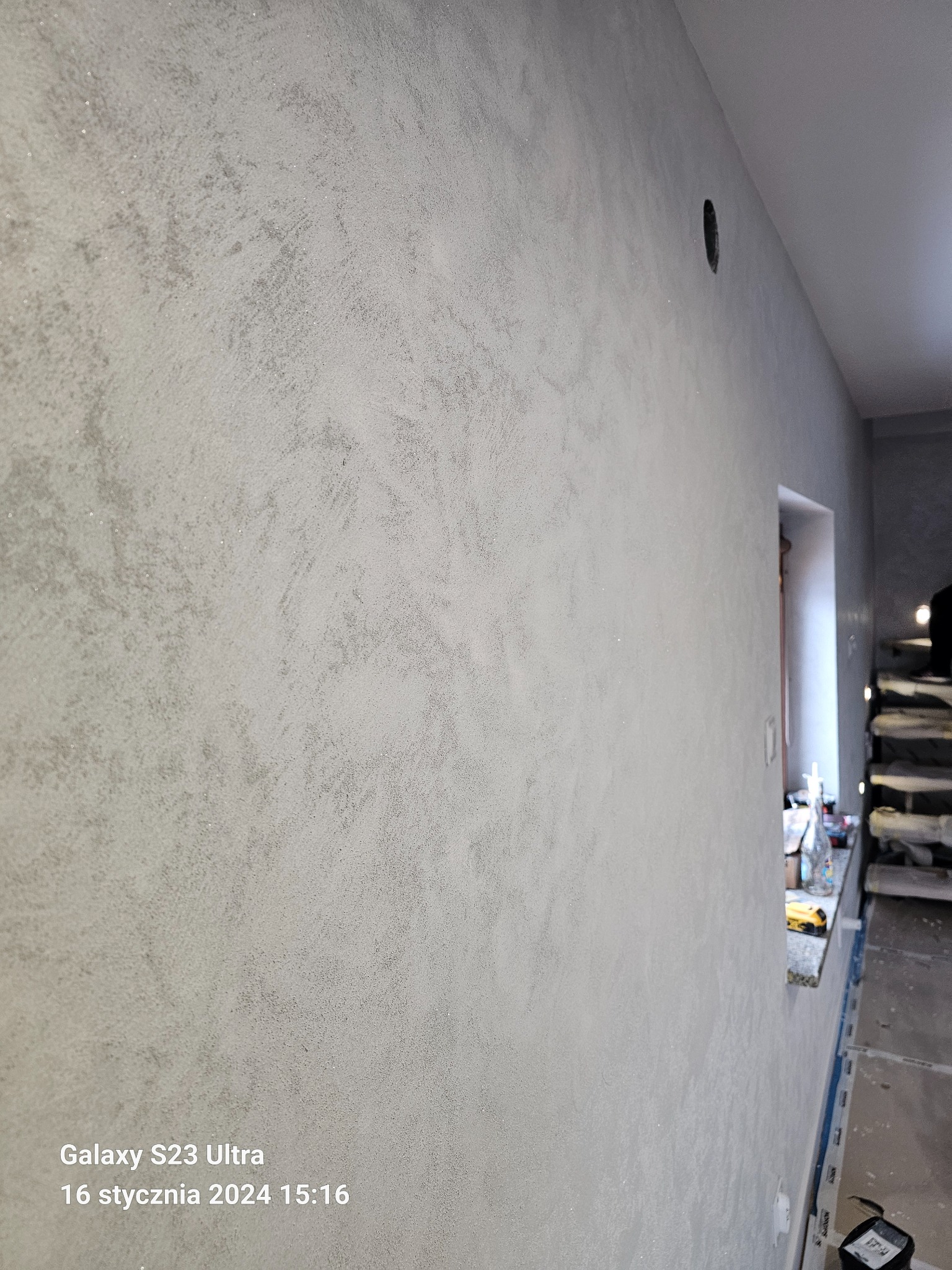 Ściana pokryta dekoracyjną, srebrzystą powłoką z widocznym otworem na gniazdko elektryczne i fragmentem okna z narzędziami w tle.