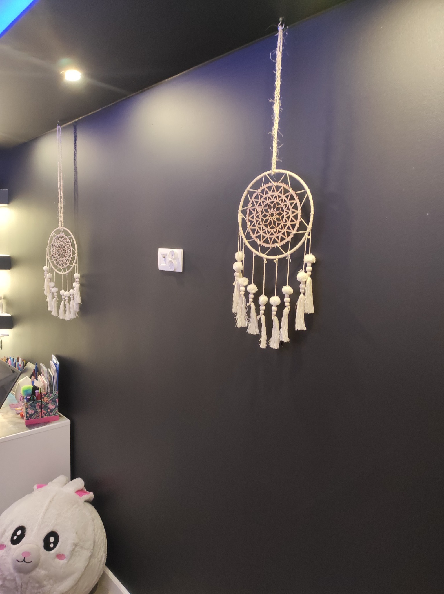 Dwa łapacze snów wiszące na pomalowanej na ciemno ścianie, obok włącznika światła, z pluszowym królikiem i biurkiem widocznymi w dolnym rogu.