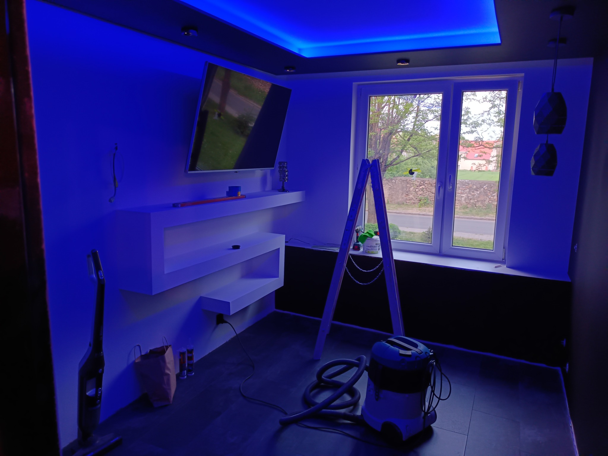 Pokój po remoncie z podświetleniem LED na suficie, drabina oparta o okno, odkurzacz i półki pod telewizorem.