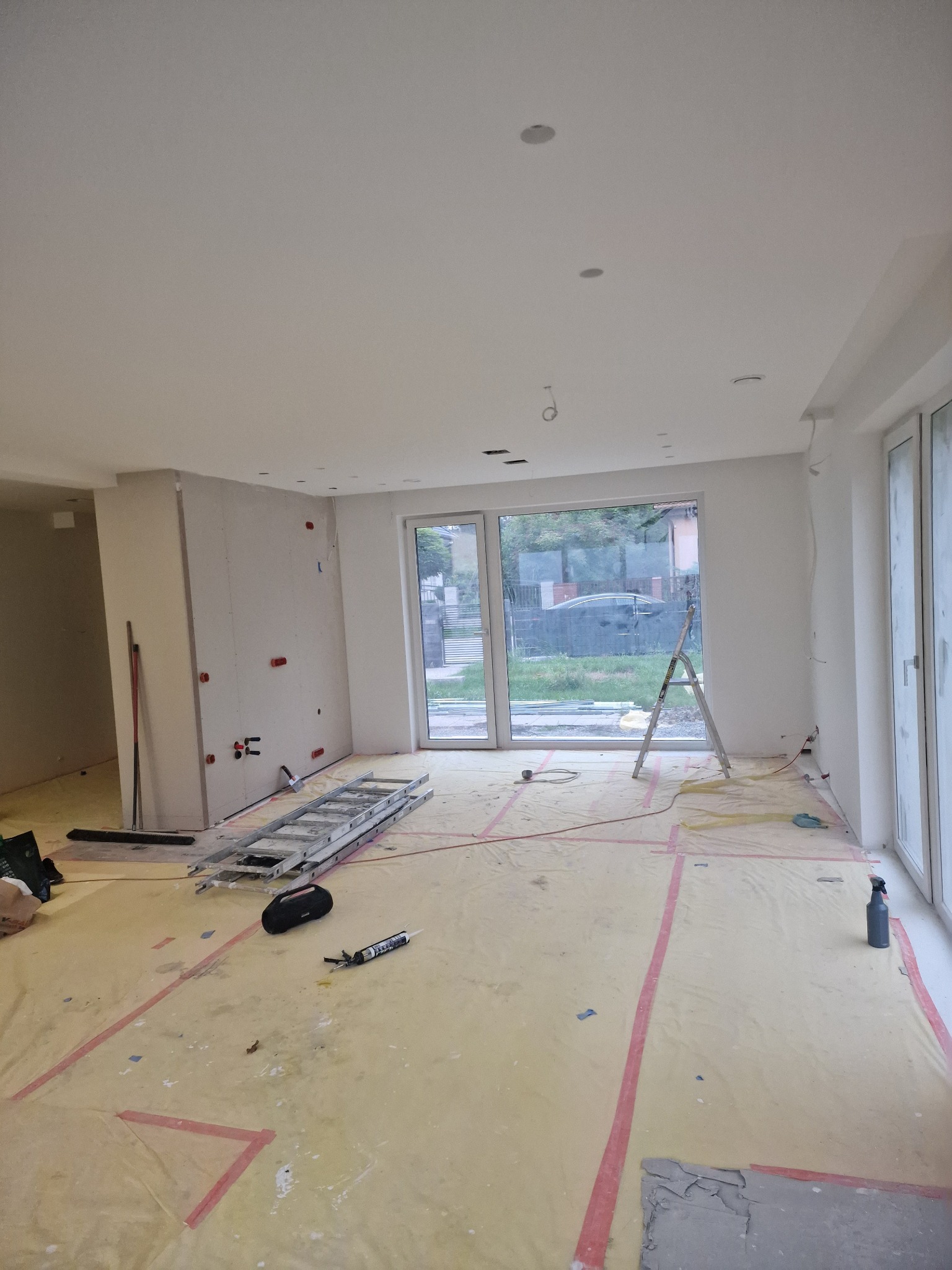 Wnętrze w trakcie prac przygotowawczych: zabezpieczona folią podłoga, ściany z płyt kartonowo-gipsowych z widocznymi instalacjami, drabina i narzędzia budowlane w przestronnym pomieszczeniu...