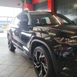 PIN UP CAR WASH SPÓŁKA Z OGRANICZONĄ ODPOWIEDZIALNOŚCIĄ - Błyszczący, czarny SUV marki Skoda Kodiaq po umyciu w myjni, widoczny z bliska na tle płytek ceramicznych i otwartej bramy wjazdowej z widokiem na inny samochód.