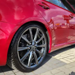 Błyszczący, czerwony Lexus IS z polakierowanymi felgami, zaparkowany na kostce brukowej w słoneczny dzień, widok z boku.