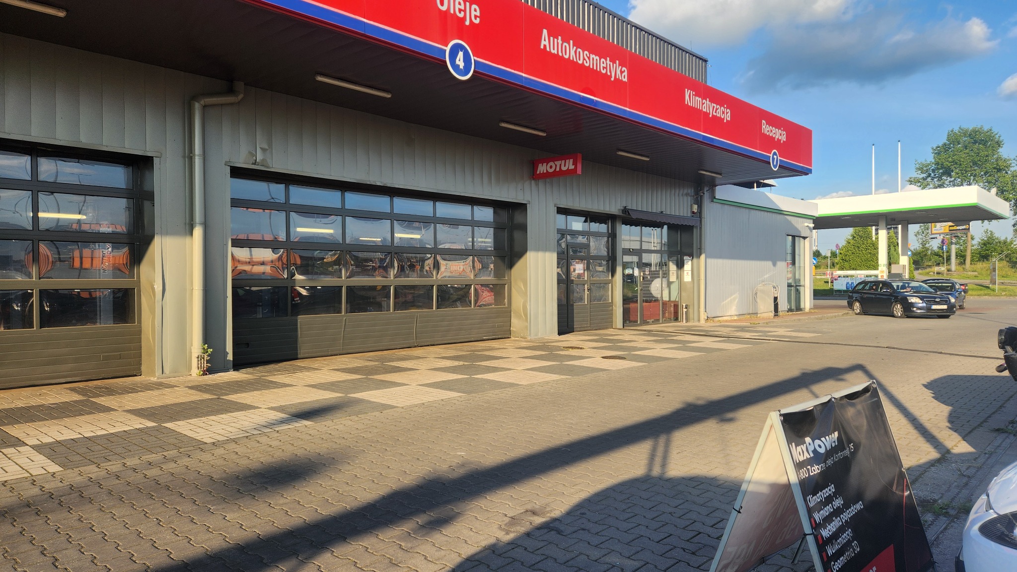 Budynek stacji obsługi pojazdów z napisem Autokosmetyka i Klimatyzacja, widoczne garaże z refleksami urządzeń w szybach, dystrybutor paliwa w tle, reklama MaxPower na chodniku.