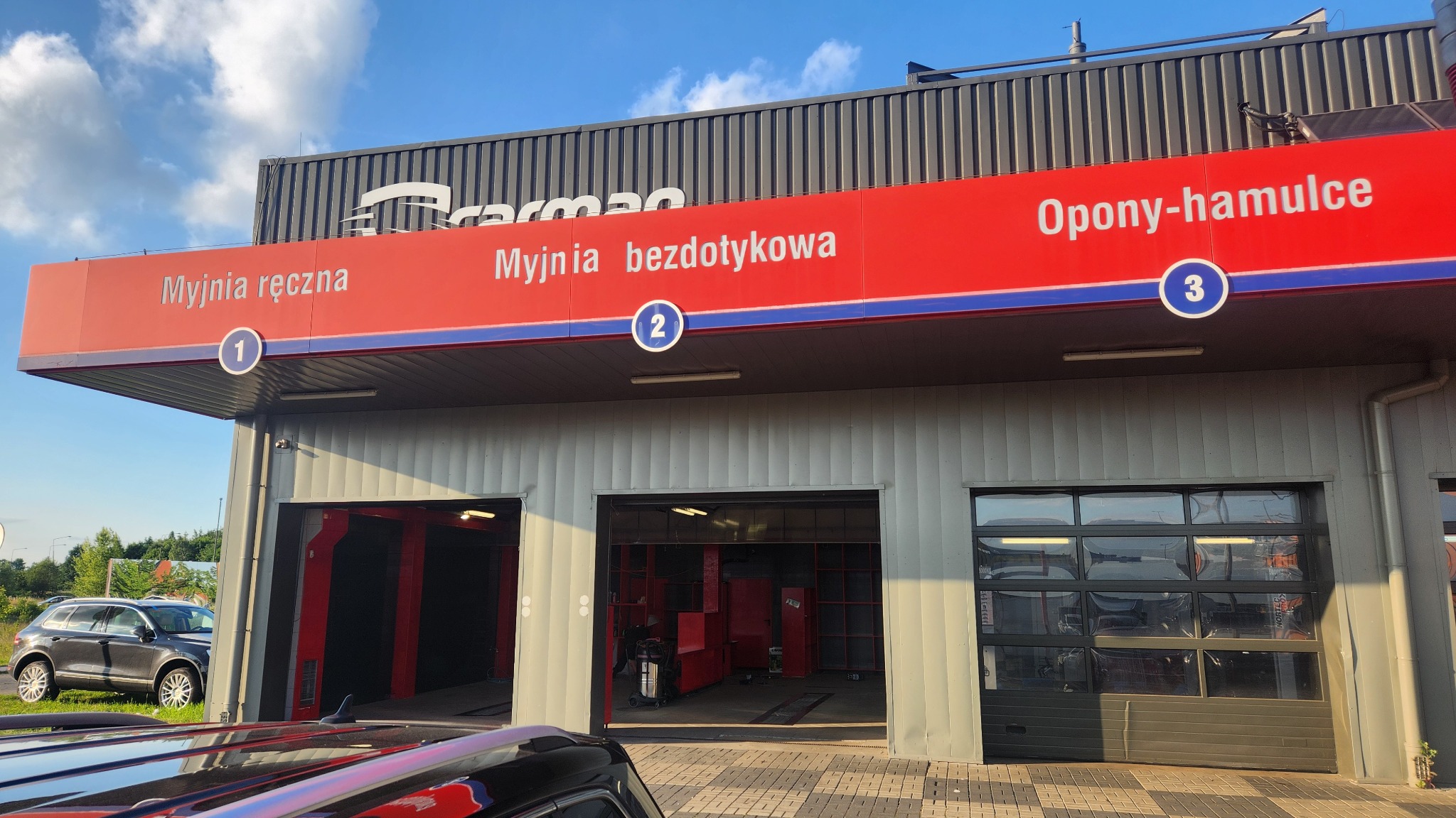 Budynek myjni samochodowej z trzema stanowiskami: myjnia ręczna, myjnia bezdotykowa i stanowisko wymiany opon, widoczne logo firmy Carman na dachu.