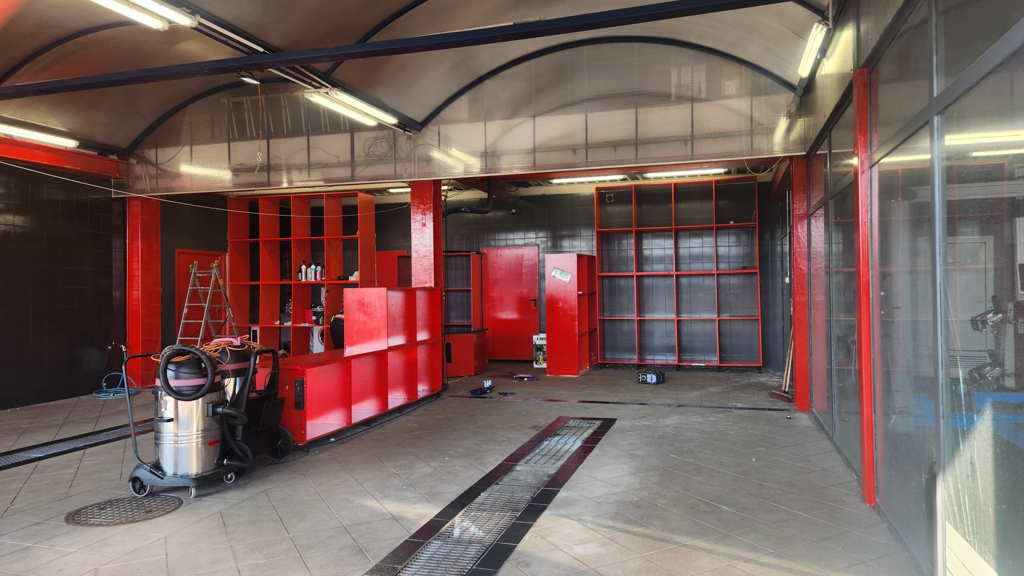 Wnętrze myjni samochodowej z widocznym odkurzaczem przemysłowym, regałami na środki czyszczące i kratką odpływową w podłodze.
