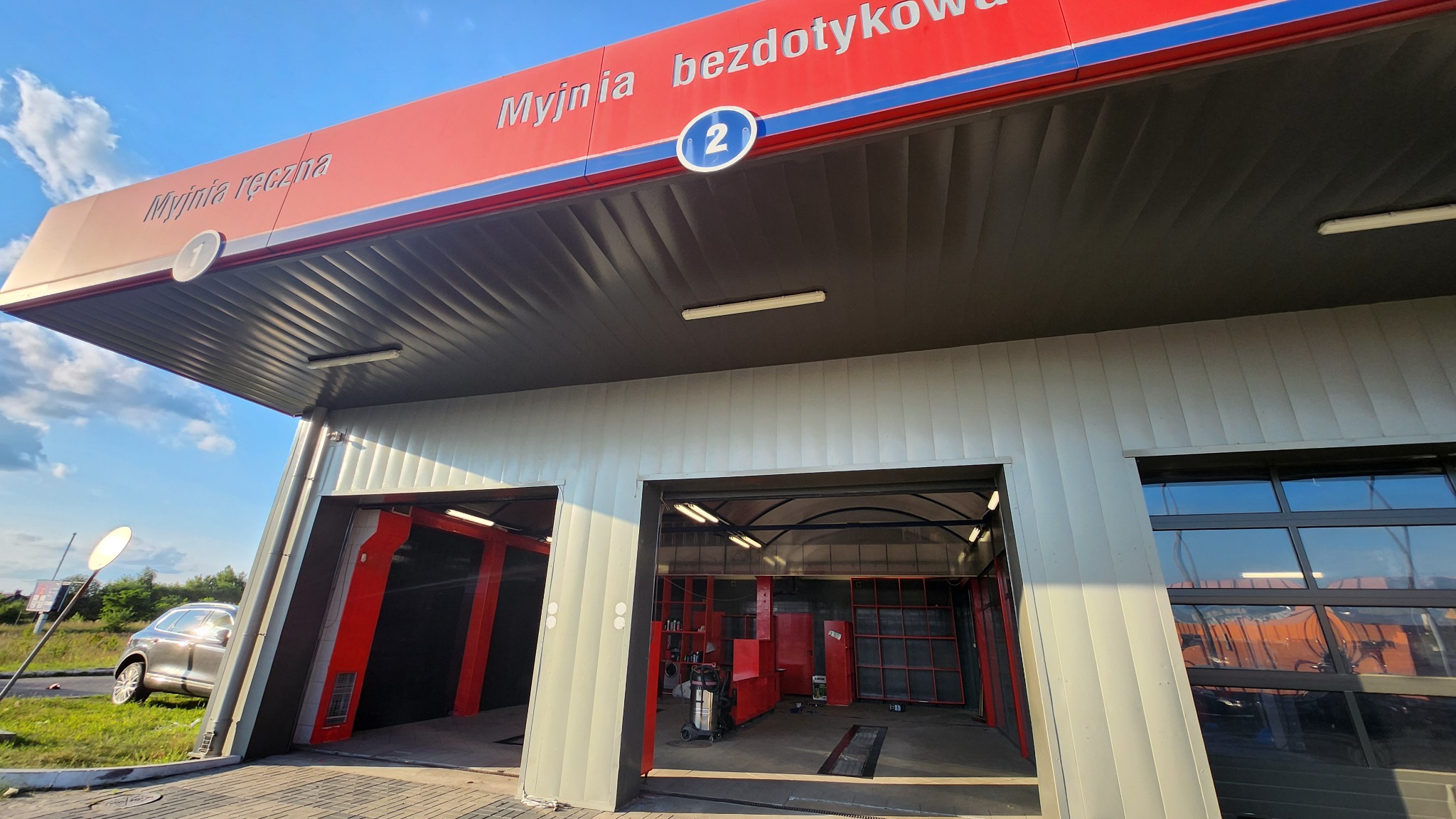 Zewnętrzny widok na dwustanowiskową myjnię bezdotykową z czerwonym daszkiem i numeracją stanowisk. Na jednym ze stanowisk widoczny wózek z urządzeniami do mycia.