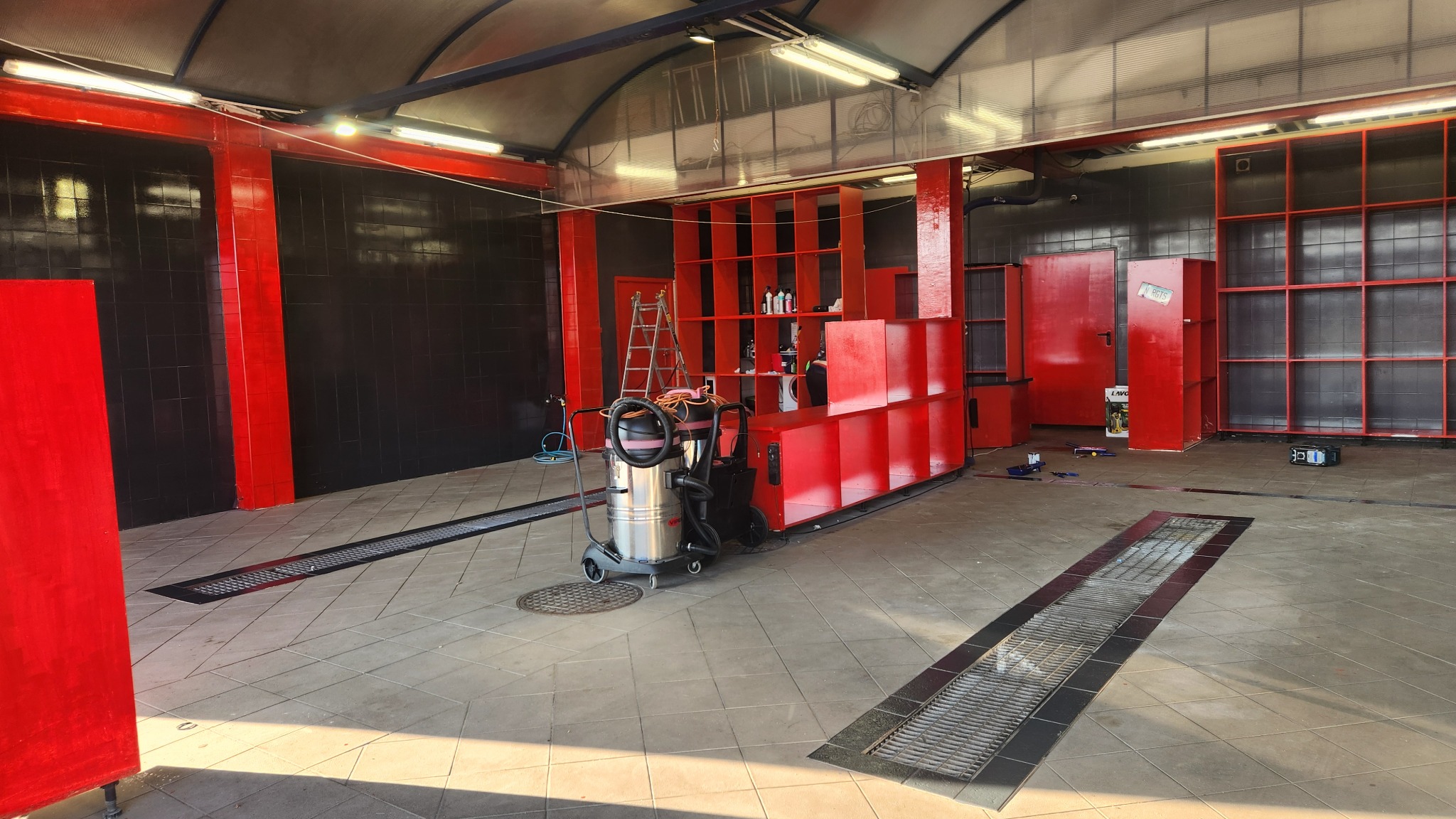 Wnętrze myjni samochodowej z odkurzaczem przemysłowym na pierwszym planie, czerwone elementy konstrukcyjne i czarne ściany, widoczne kratki odpływowe w podłodze.