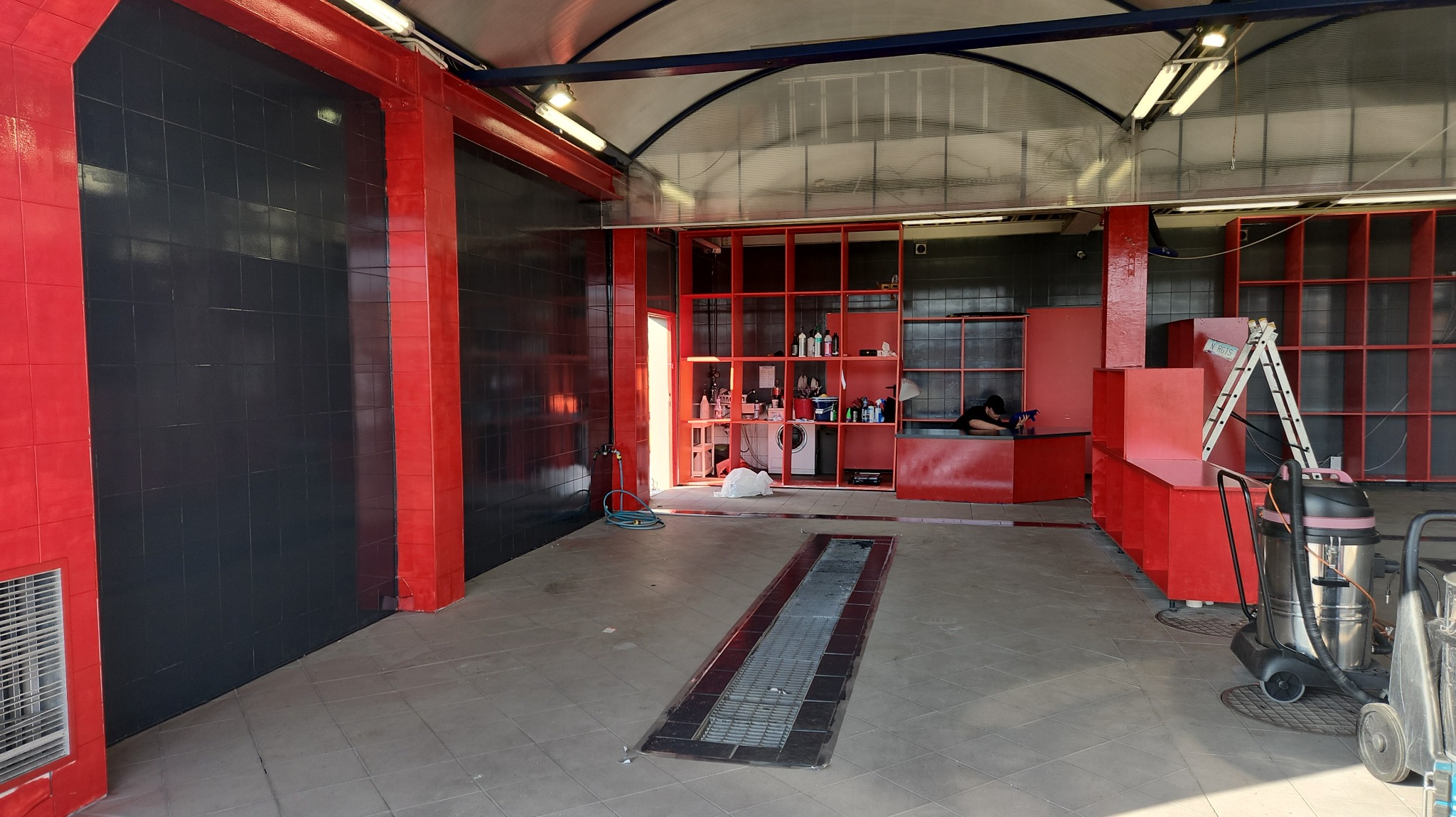 Wnętrze myjni samochodowej z czarno-czerwonymi elementami, widoczny kanał odpływowy w podłodze, regał z preparatami, pralka i odkurzacz przemysłowy.
