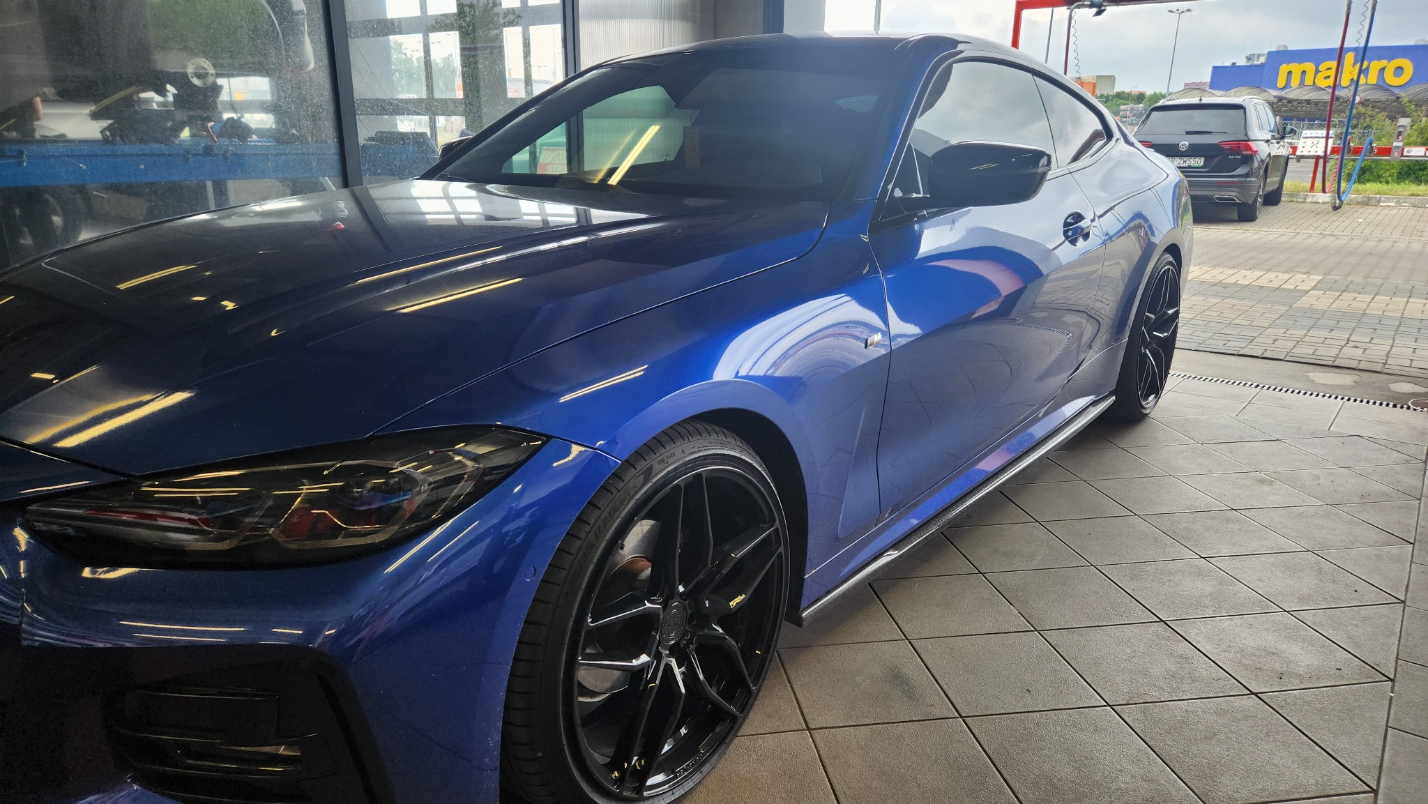 Błyszczący, granatowy samochód coupe po umyciu, z widocznymi kroplami wody, czarnymi felgami i odbiciami światła na lakierze, zaparkowany na kafelkowanej posadzce myjni.