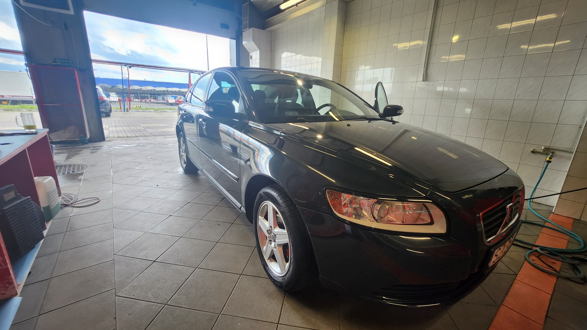 Czarny samochód Volvo S40 po umyciu w myjni, widoczne krople wody na karoserii, otwarte drzwi kierowcy, płytki na ścianach i posadzce.