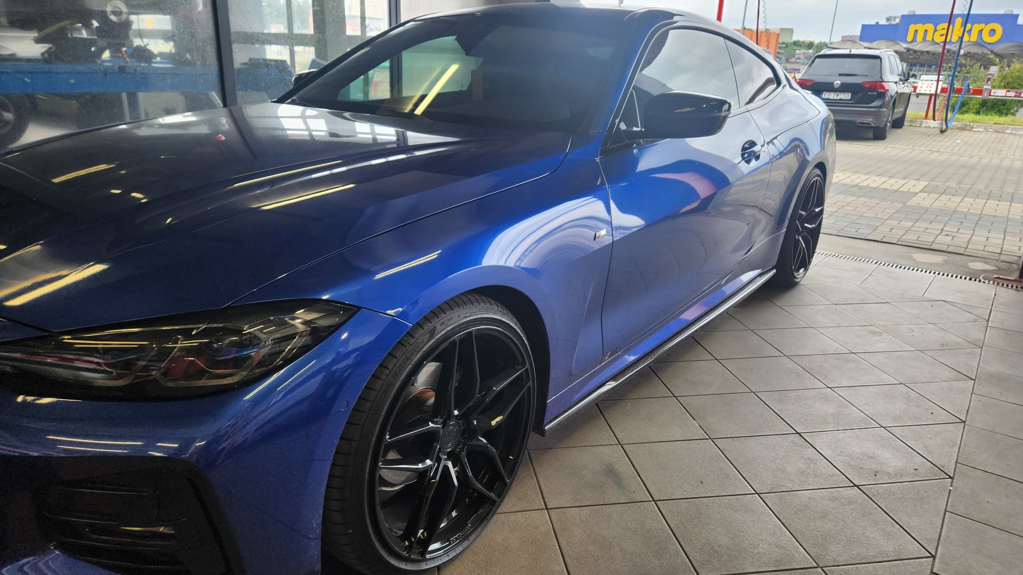 Błyszczący, granatowy samochód coupe na stanowisku myjni z czarnymi felgami, widoczny inny samochód w tle i budynek Makro.