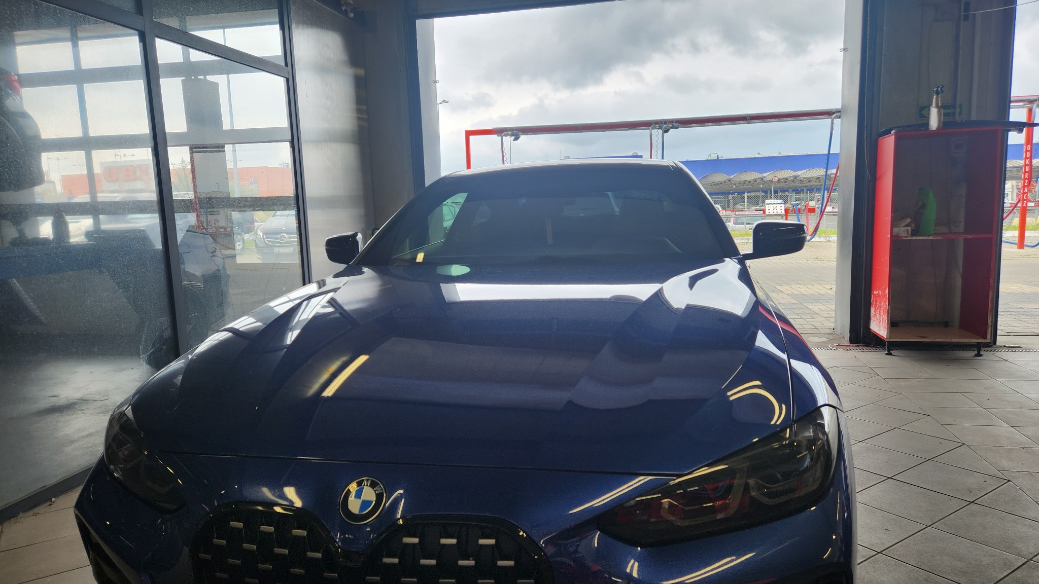 Granatowy samochód BMW, widok z przodu, wewnątrz myjni automatycznej z widocznymi elementami konstrukcji myjącej i otwartą bramą wjazdową.