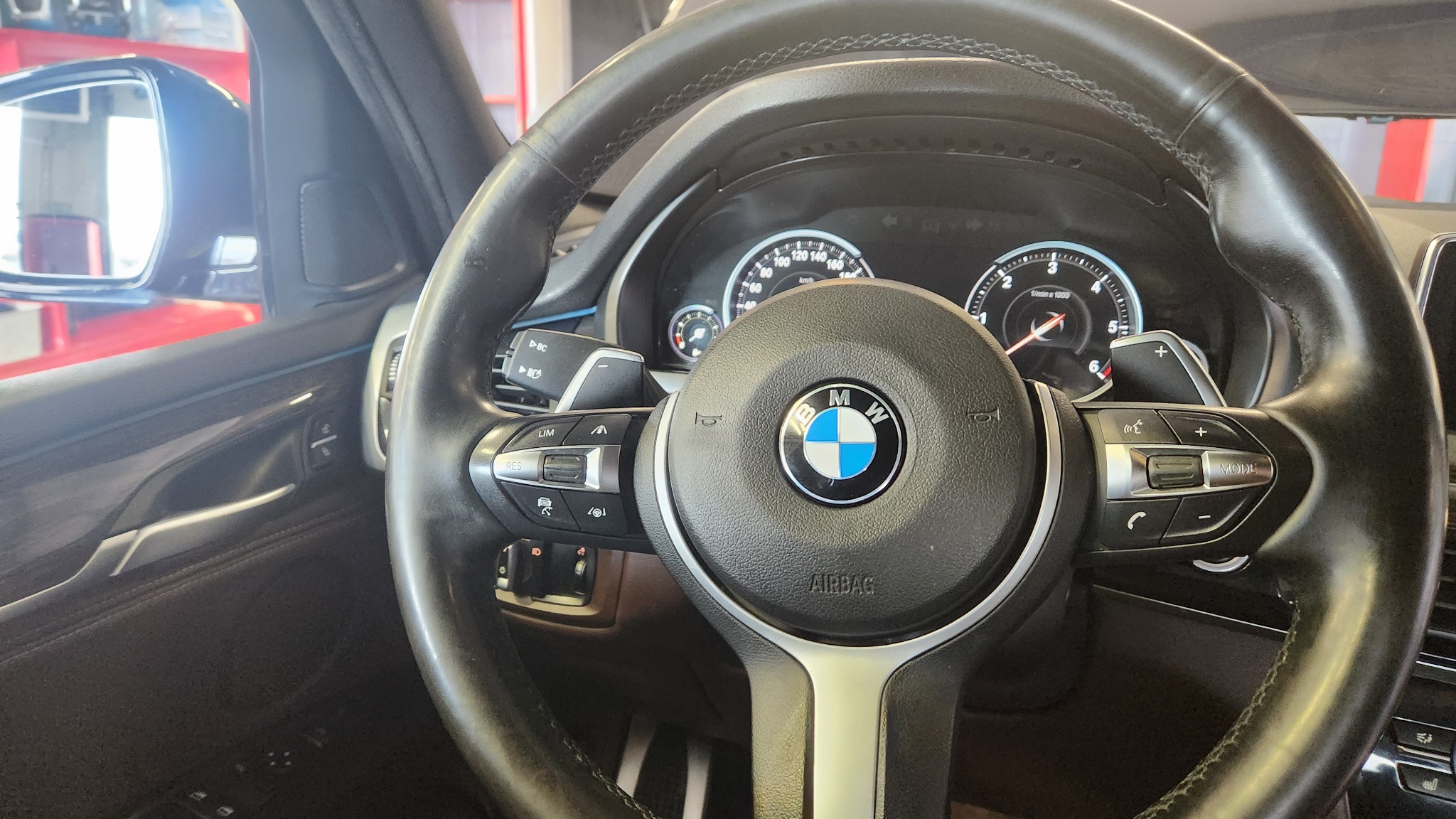 Czarna skórzana kierownica BMW z widocznym logo, deską rozdzielczą i elementami sterowania, wnętrze samochodu z czerwonym fotelem widocznym w lusterku.