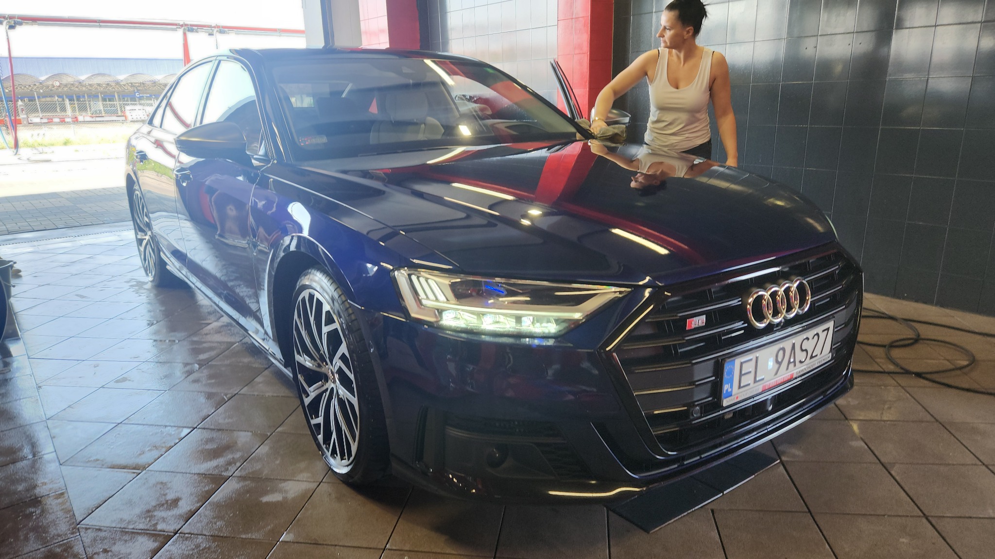 Granatowy samochód Audi A8 po umyciu, widoczna kobieta wycierająca karoserię ściereczką, mokra posadzka i oświetlenie hali.