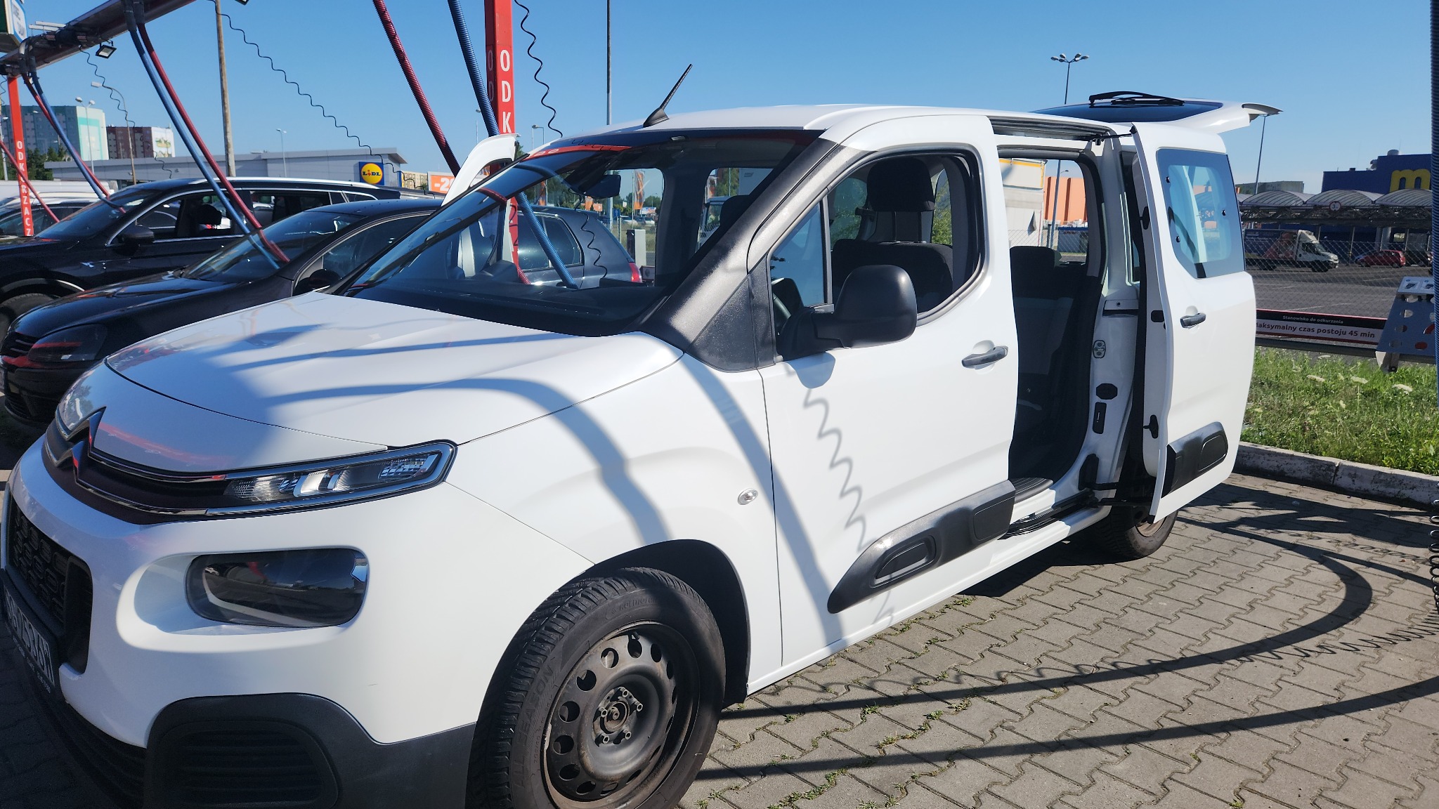 Biały samochód dostawczy z otwartymi drzwiami, stojący na kostce brukowej przed myjnią samochodową w słoneczny dzień.