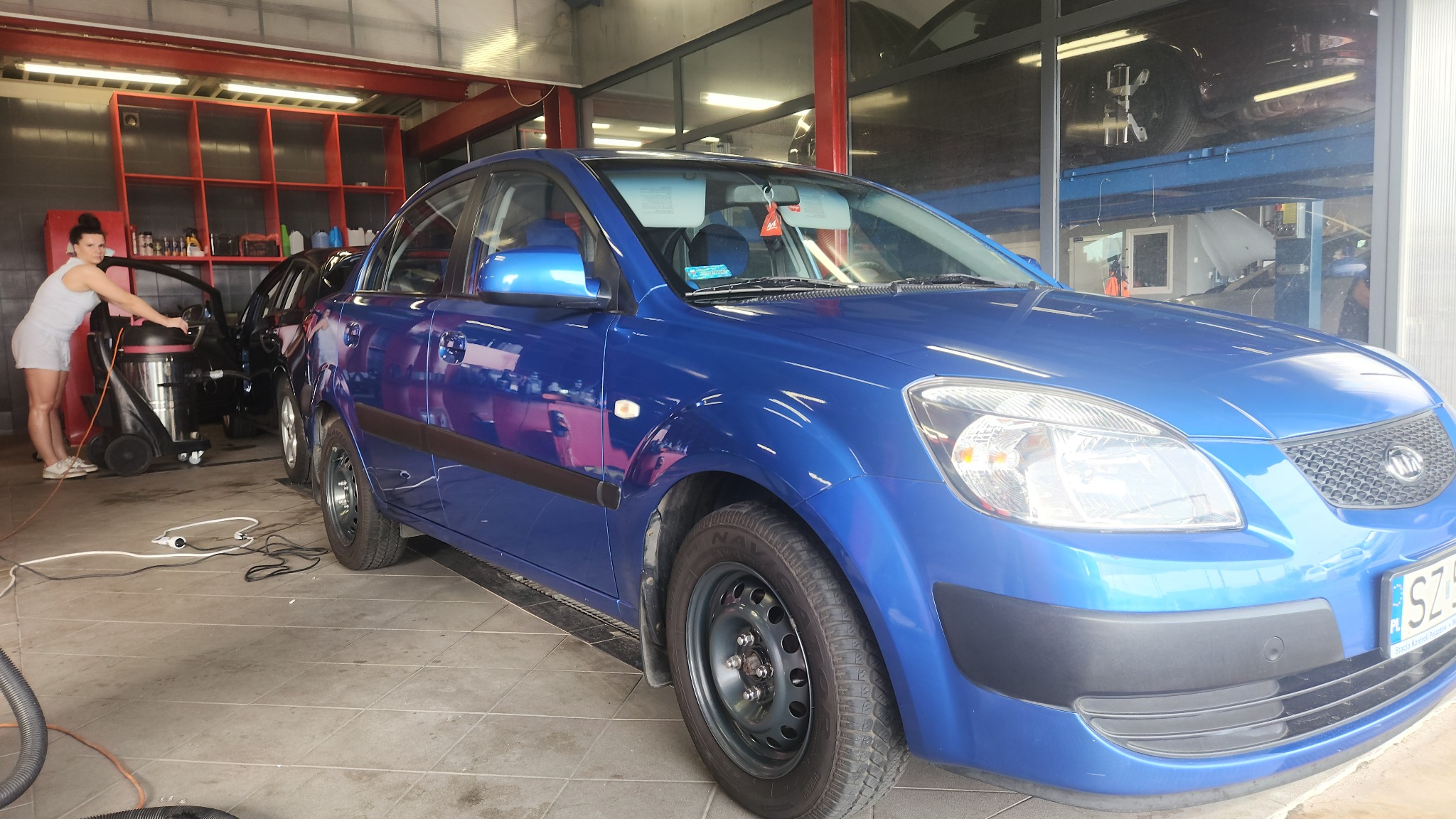 Wnętrze myjni samochodowej: niebieska Kia Rio z widoczną tablicą rejestracyjną w trakcie czyszczenia, obok pracownica z odkurzaczem piorącym i czarnym samochodem w tle, na podnośniku widoczny inny...