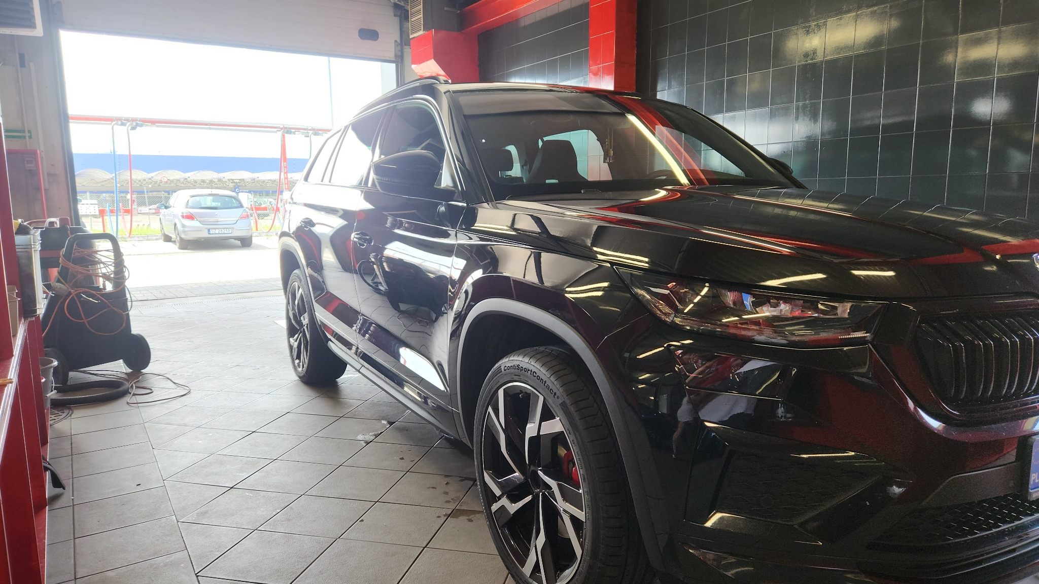 Błyszczący, czarny SUV marki Skoda Kodiaq po umyciu w myjni, widoczny z bliska na tle płytek ceramicznych i otwartej bramy wjazdowej z widokiem na inny samochód.