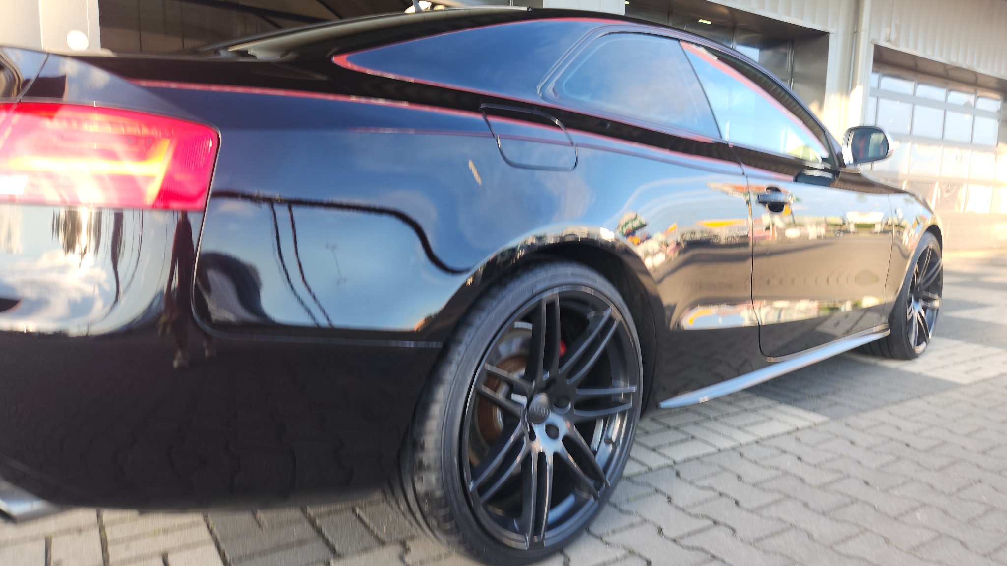 Błyszczący, czarny samochód sportowy Audi z czarnymi felgami i czerwonymi akcentami, zaparkowany na kostce brukowej, widok z tyłu i boku.