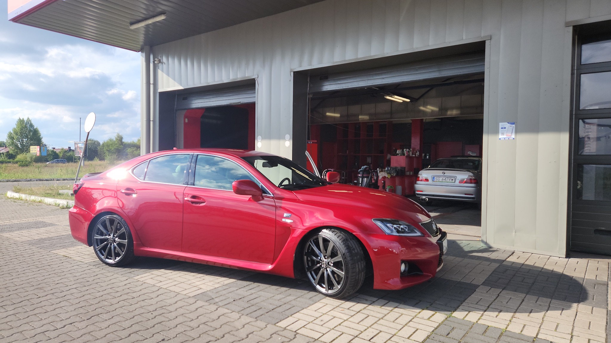 Błyszczący, czerwony Lexus IS-F po umyciu, zaparkowany przed wjazdem do myjni z otwartą bramą i widocznym drugim samochodem w środku.