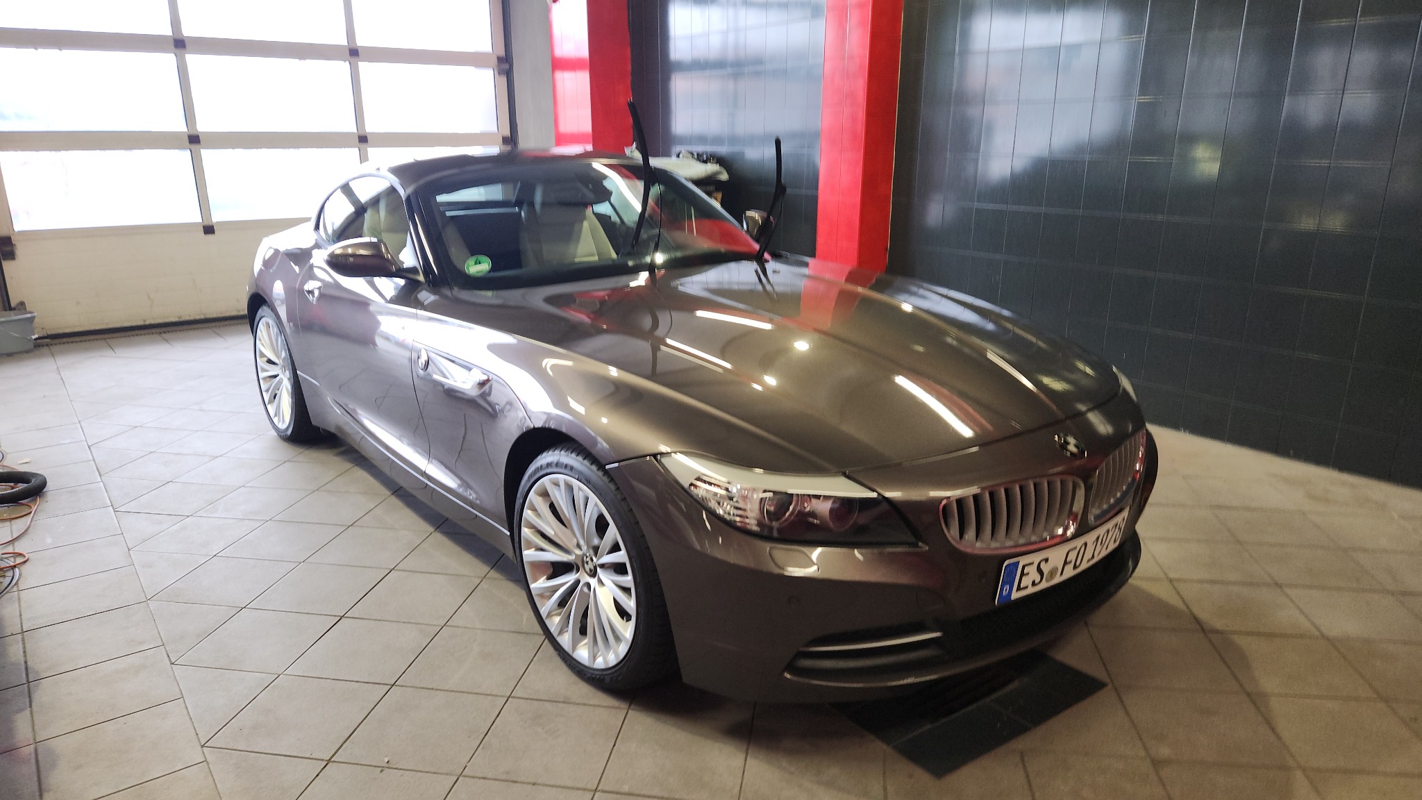 Brązowy kabriolet BMW Z4 po umyciu, z widocznymi kroplami wody na karoserii, stojący w jasnym pomieszczeniu z kafelkami na podłodze i pionowymi panelami na ścianie.