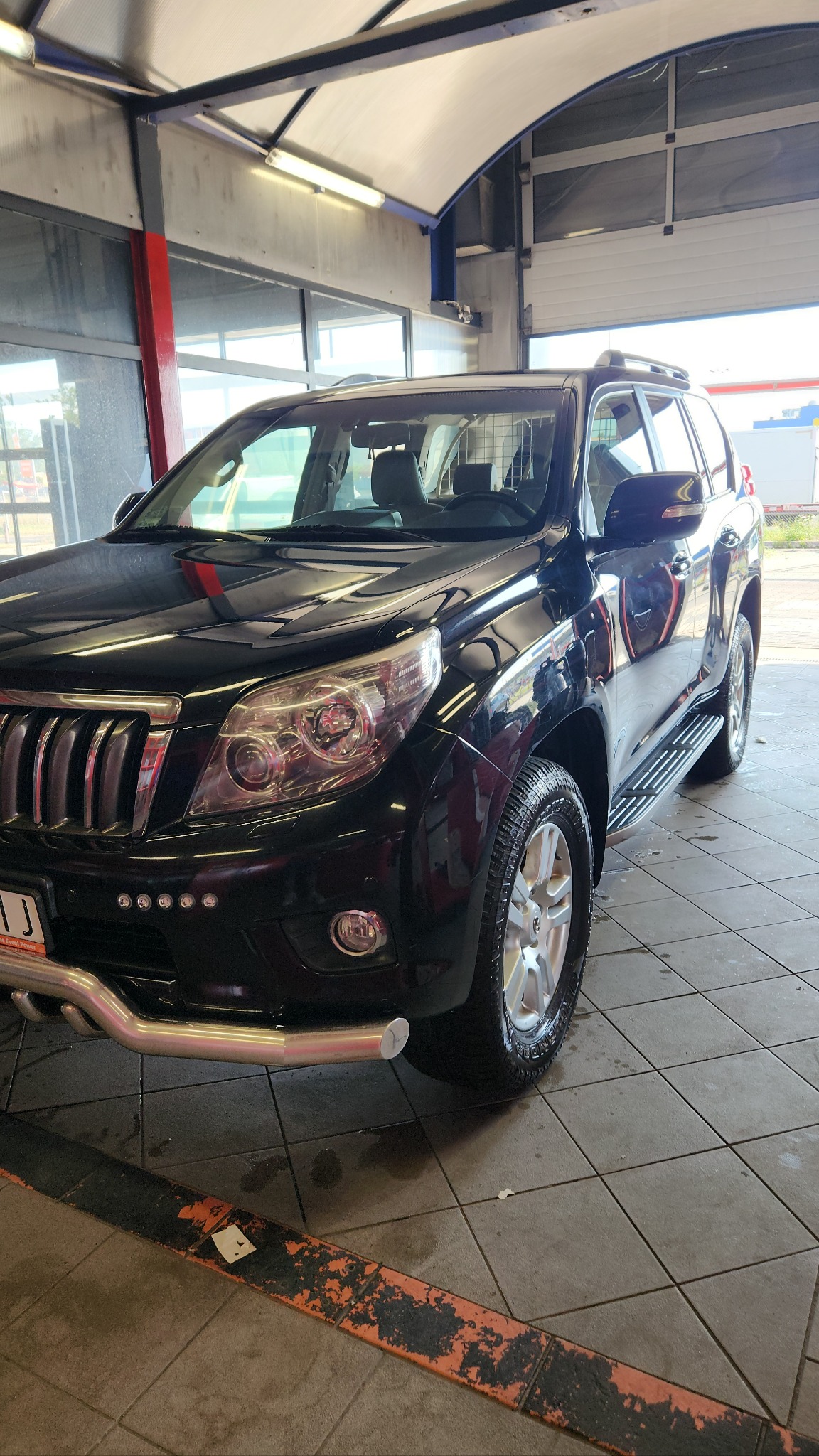 Czarny SUV Toyota Land Cruiser po myciu w myjni automatycznej, widoczne krople wody na karoserii i oponach, metalowy orurowanie przedniego zderzaka.