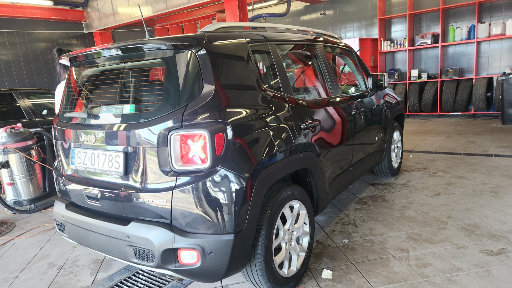 Czarny Jeep Renegade Limited, widok z tyłu, w trakcie czyszczenia w myjni; widoczny odkurzacz przemysłowy i mokra podłoga.